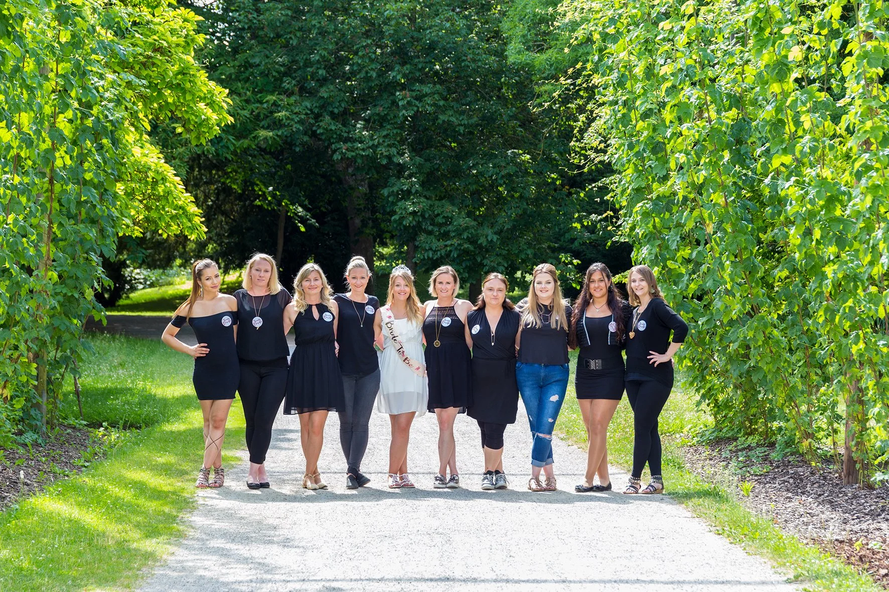 Gruppe von Frauen am Tag der Abiturfeier, beim Spaziergang im Park, alle tragen schwarze Kleider oder Outfits, die Sonne scheint, im Hintergrund Bäume und grünes Laub.