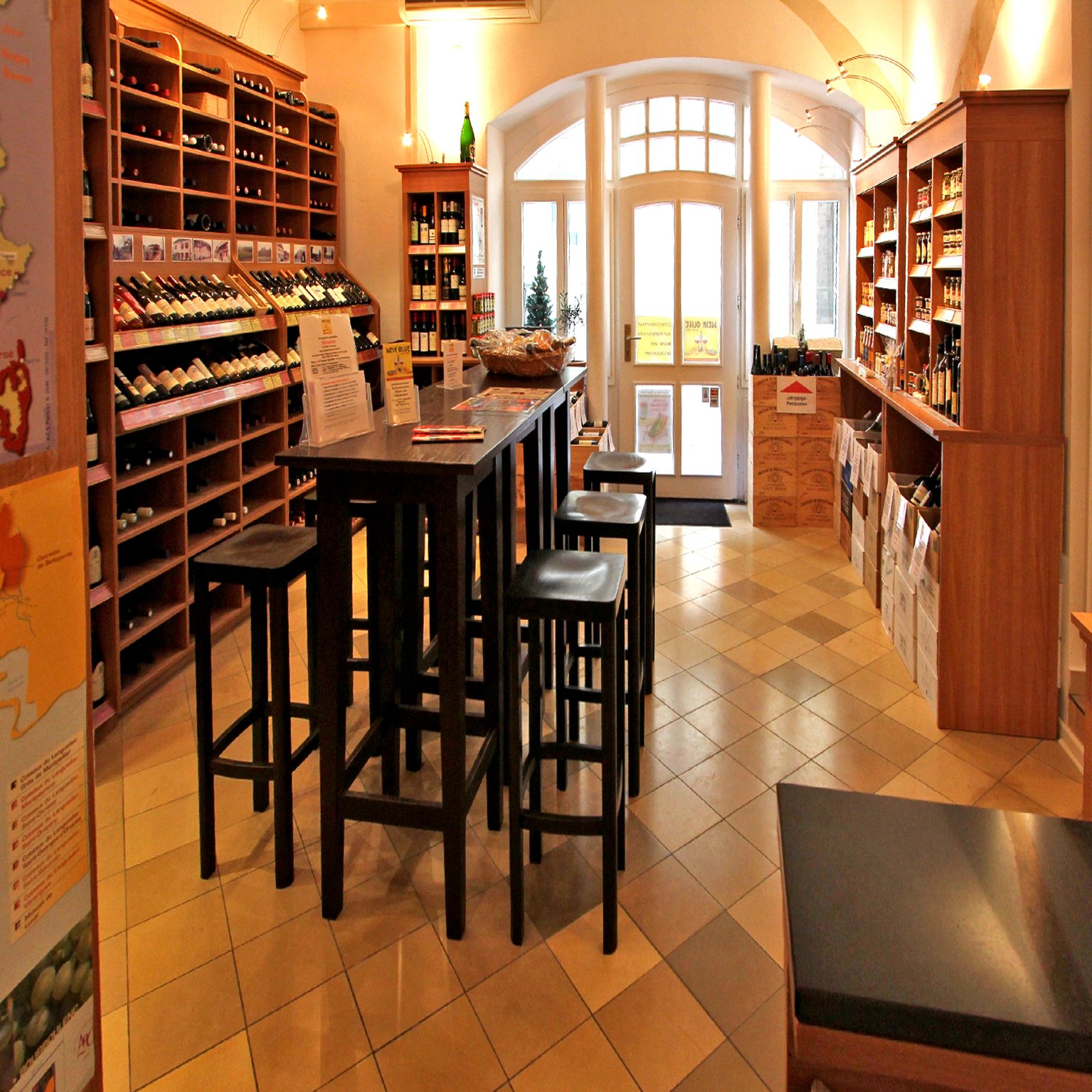 Innenaufnahme eines Weingeschäfts mit Holzregalen voller Weinflaschen, einem hohen Stehtisch mit Barhockern, und einem Eingang mit GlasTür im Hintergrund.