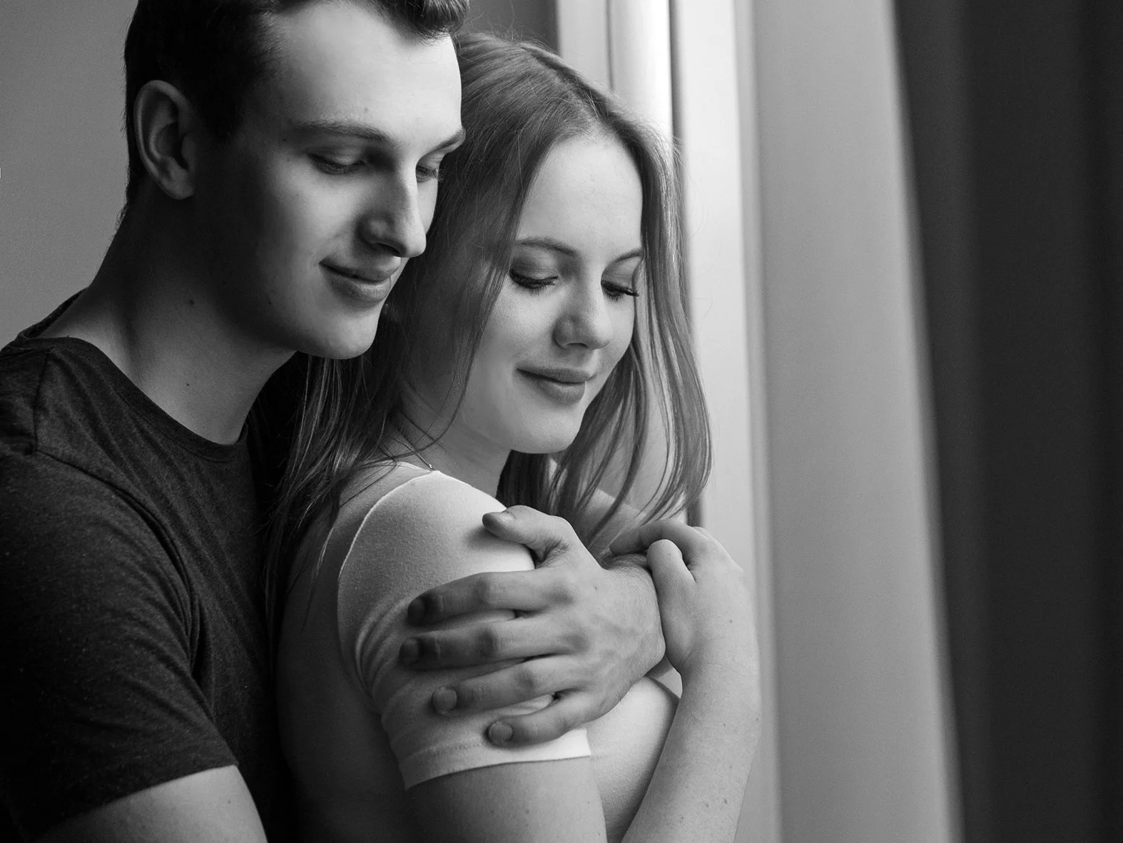 Ein Mann hält eine Frau eng umarmt, beide mit geschlossenen Augen und einem Lächeln. Schwarz-weiß-Fotografie, Gefühl von Nähe und Zuneigung.