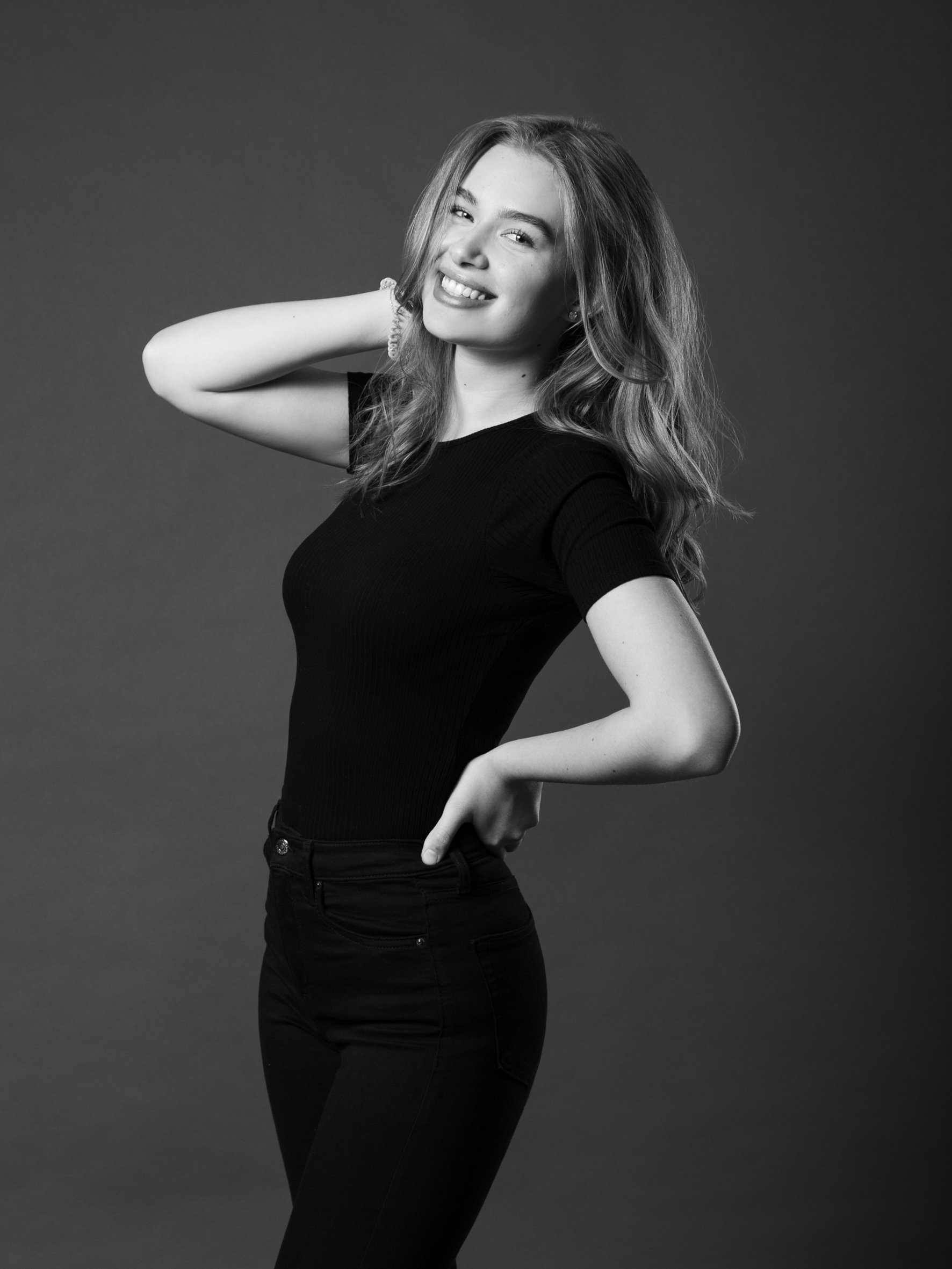 Schwarz-weiß Foto eines lachenden jungen Frau in Schwarz mit lockigem Haar vor einfachem dunklem Hintergrund.