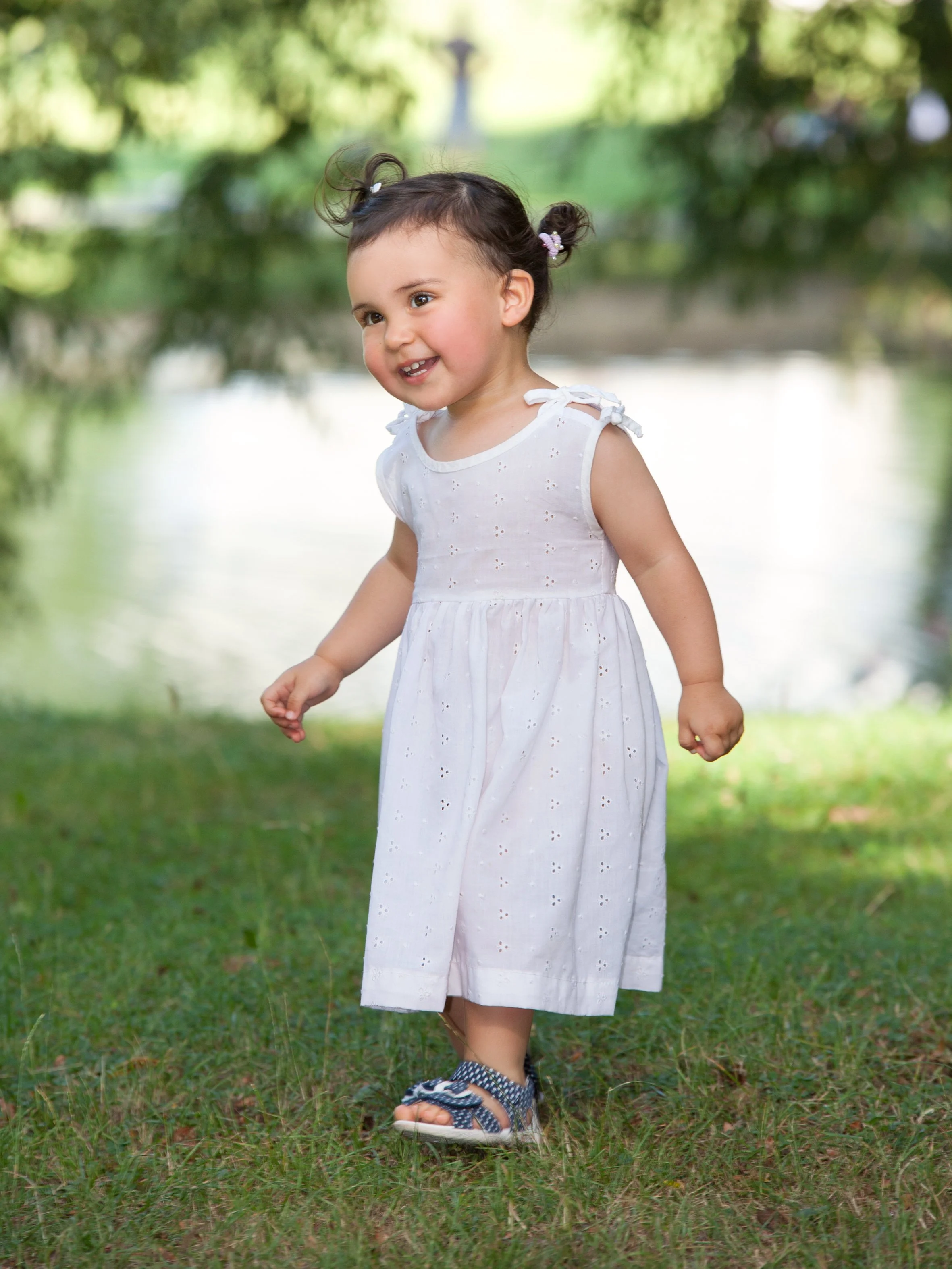 Ein kleines Mädchen läuft glücklich im Freien entlang eines Flusses, trägt ein weißes Kleid und Sandalen.