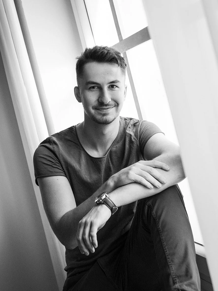 Schwarz-weiß-Foto eines jungen Mannes sitzt auf dem Boden neben einem Fenster, lächelt, trägt ein T-Shirt und eine Armbanduhr.