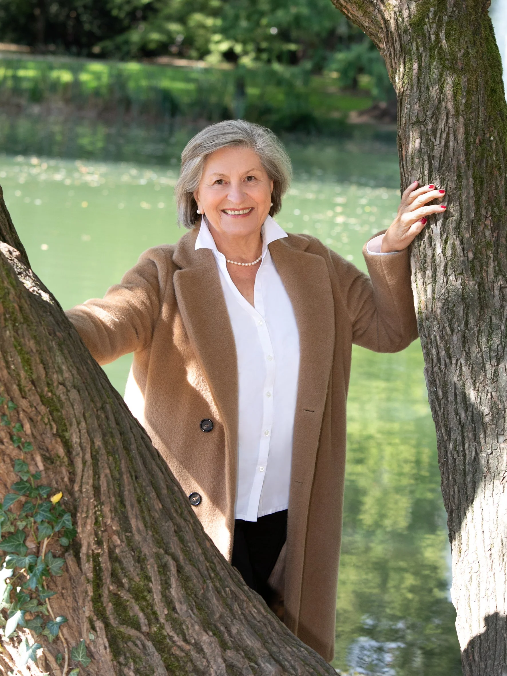 Eine ältere Frau mit grauem Haar, die ein braunes Mantel trägt, steht zwischen zwei Baumstämmen an einem Teich und lächelt in die Kamera.