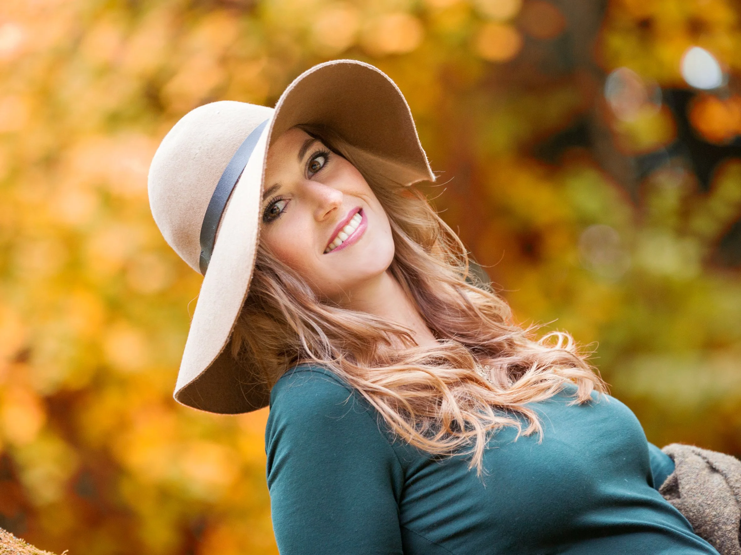 Eine lachende junge Frau trägt einen großen weichen Strohhut mit einem schwarzen Band und ein dunkles T-Shirt, während sie im Herbst vor einer Baumlandschaft steht.