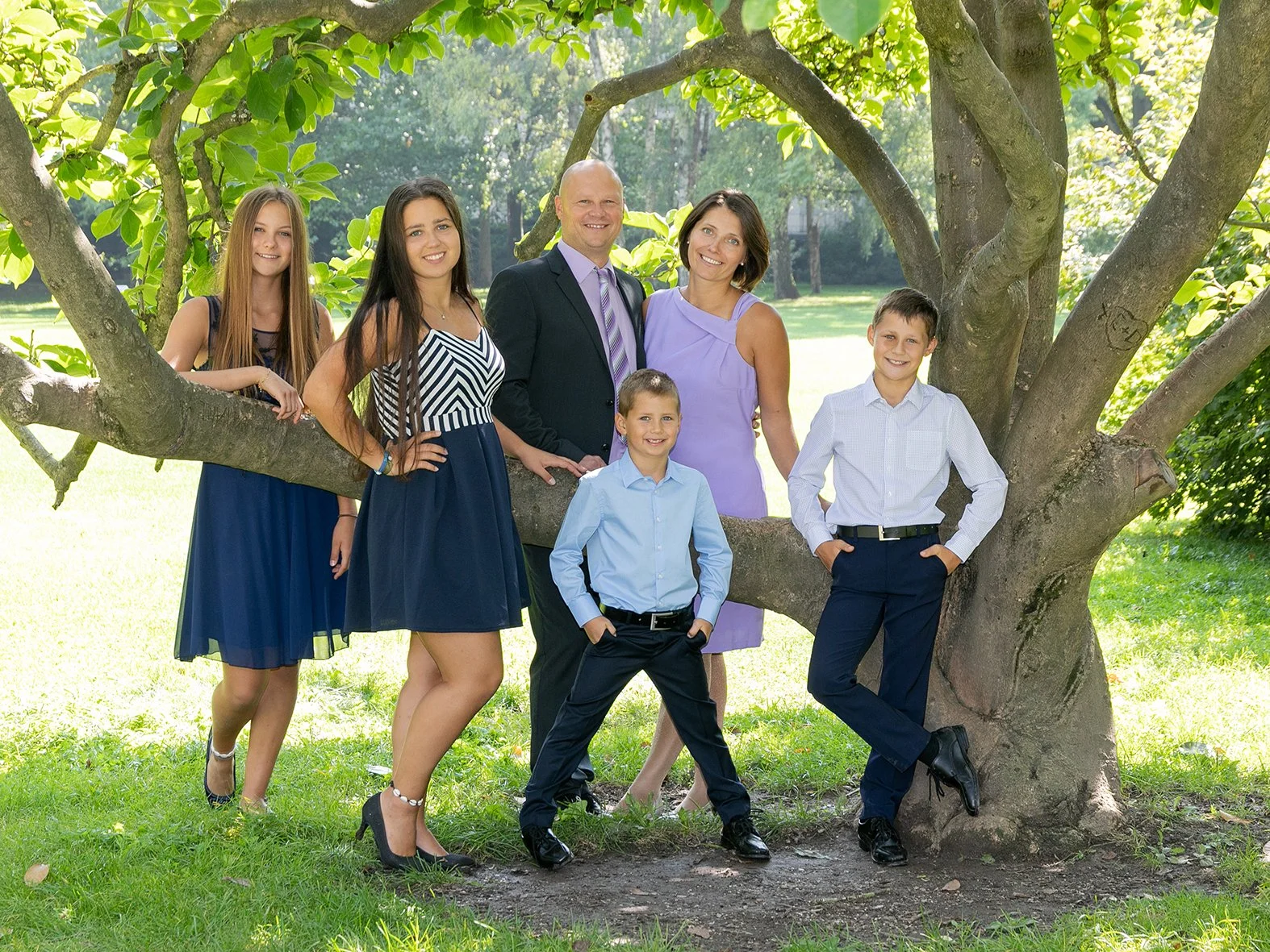 Familie posiert im Park vor einem Baum, Kinder und Erwachsene, alle lächelnd, in formeller Kleidung, sonniger Tag, grüne Umgebung.