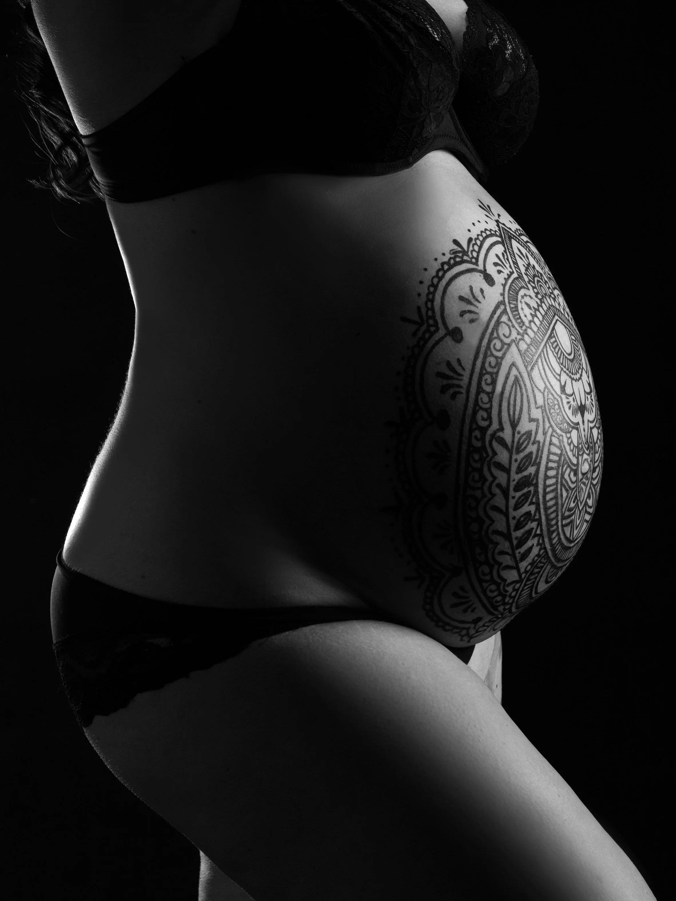 Nahaufnahme des schwangeren Bauches einer Frau mit Tattoo, Wearings Lingerie, schwarzer Hintergrund, schwarzweißer Kontrast.