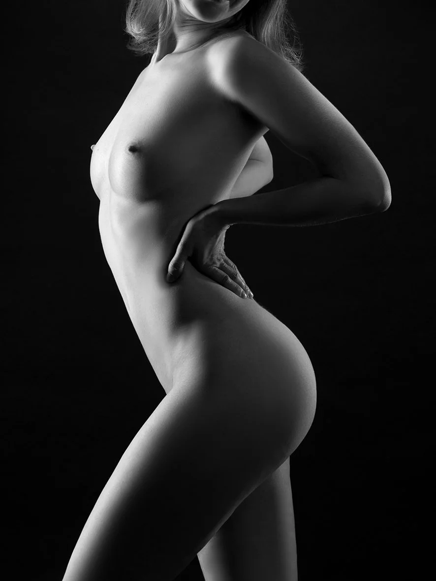 Schwarz-Weiß-Fotografie einer nackten Frau in leichter Seitenansicht, mit einer Hand auf der Taille, dunklem Hintergrund.
