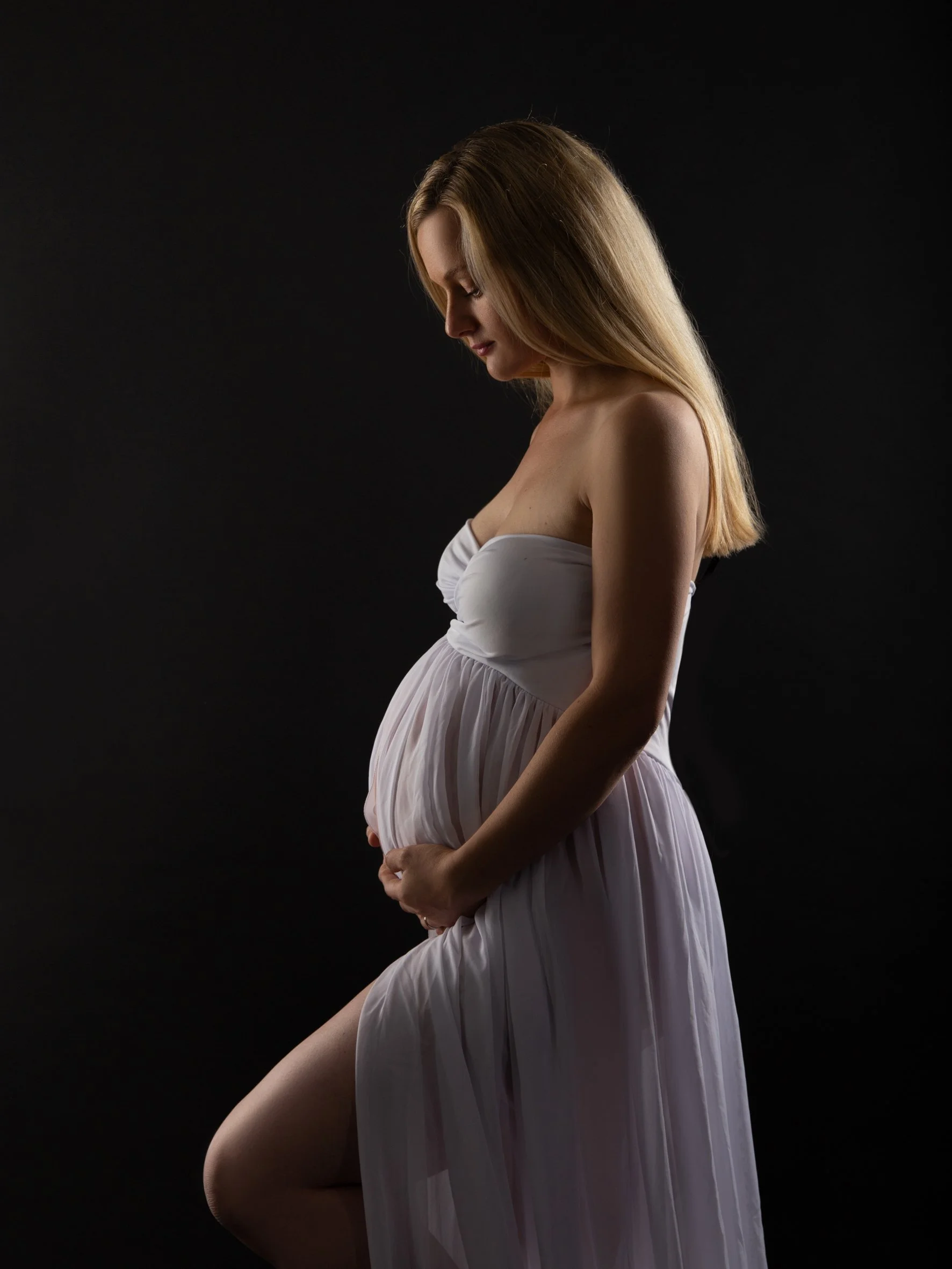 Schwangere Frau in weißem Kleid, seitlich betrachtet, vor schwarzem Hintergrund.