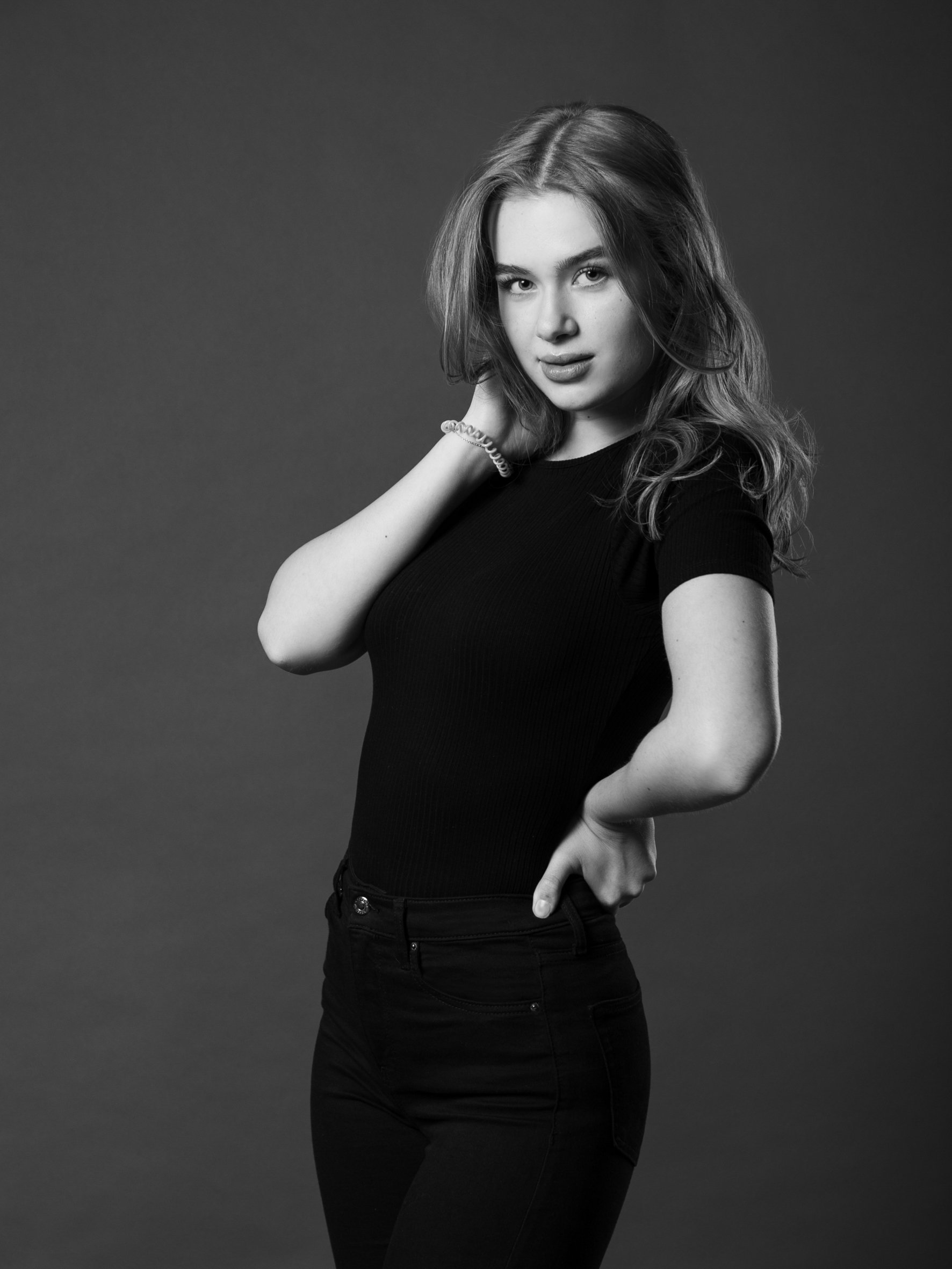 Schwarz-Weiß-Foto eines jungen Mädchens in schwarzer Kleidung, das an den Haaren zieht, mit einer selbstbewussten Pose, vor einem dunklen Hintergrund.