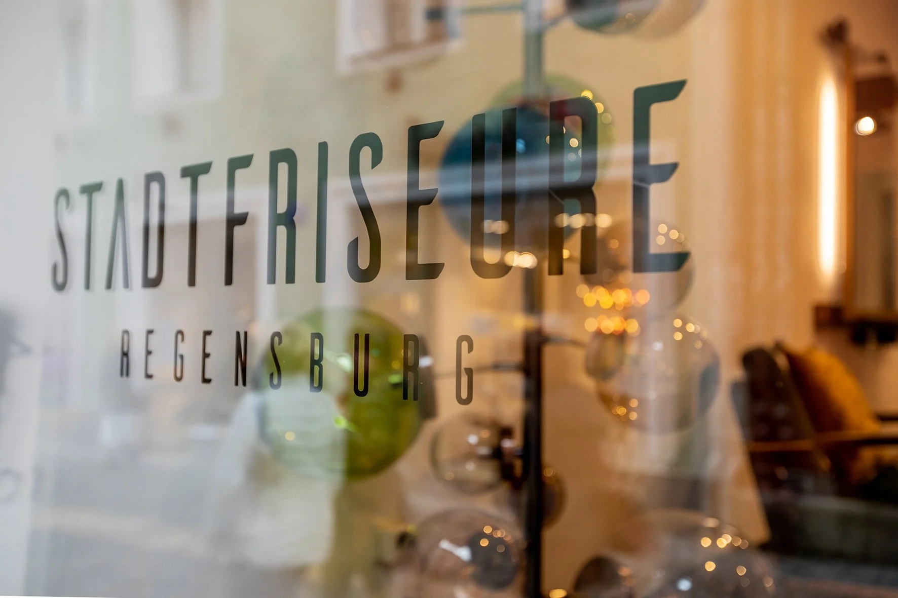 Blick durch eine gläserne Tür mit Aufschrift 'Stadteresse Regensburg', im Hintergrund sind ausgefallene Lampen und eine gemütliche Sitzcouch.