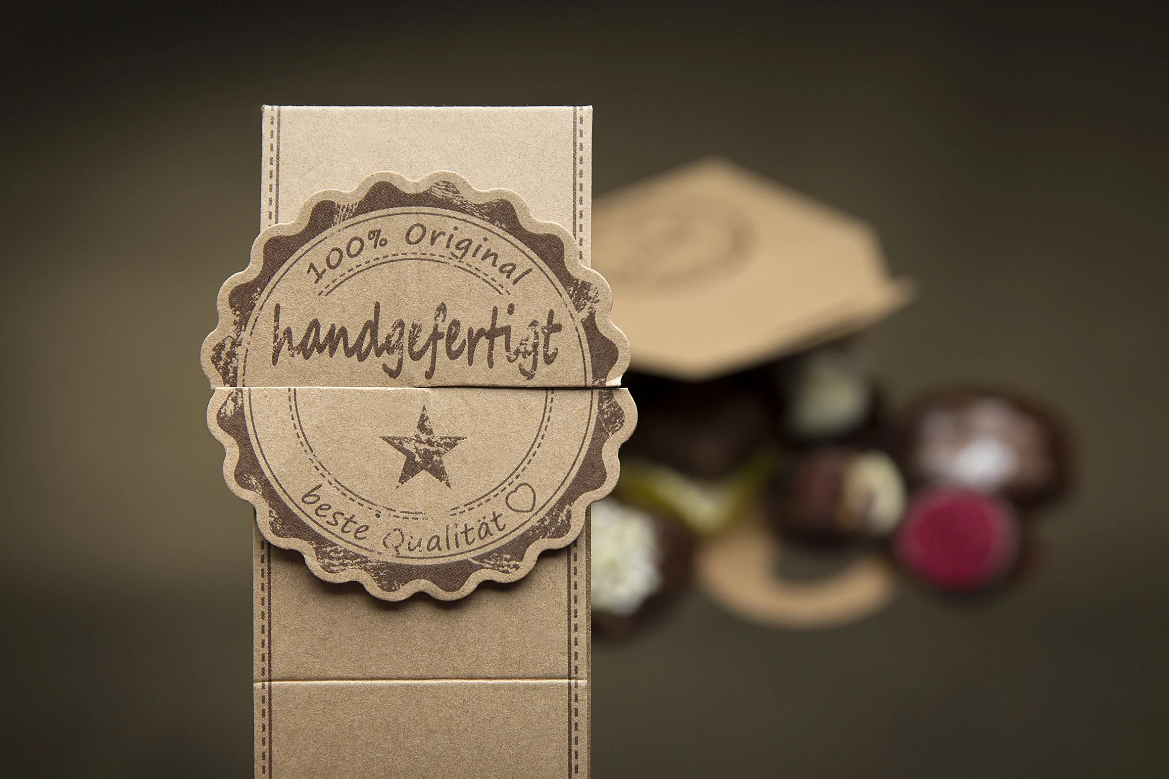 Nahaufnahme eines braunen Gewürz- oder Kakaoverpackung mit einem runden Siegel, das '100% Original handgefertigt, beste Qualität' sagt, im Hintergrund sind verschiedene hörnchenartige Süßigkeiten zu sehen.