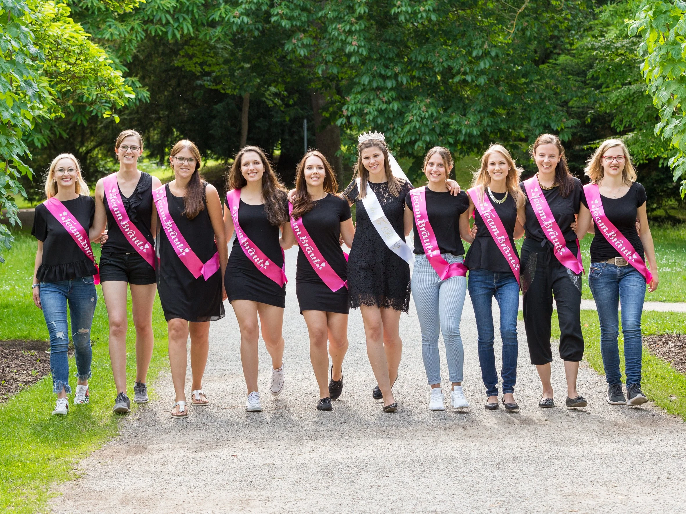 Junge Frauen bei einer Abschlussfeier im Park, alle tragen schwarze Kleidung und pinke Schärpen mit der Aufschrift 'Mein Abschluss!', eine Frau trägt eine Krone und einen weißen Schatz. Sie lachen und haben die Arme um die Schultern.