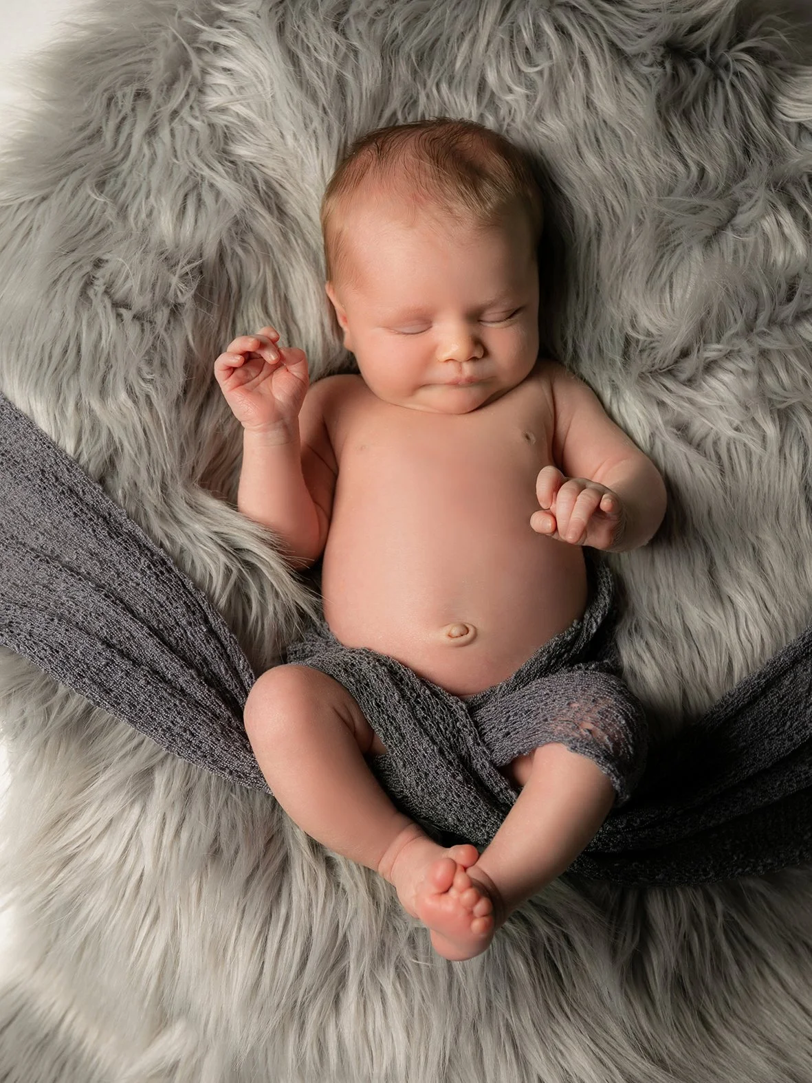 Ein schlafender Baby liegt auf einem kuscheligen grauen Fellkissen, halb in einer grauen Stoffhose eingepackt.