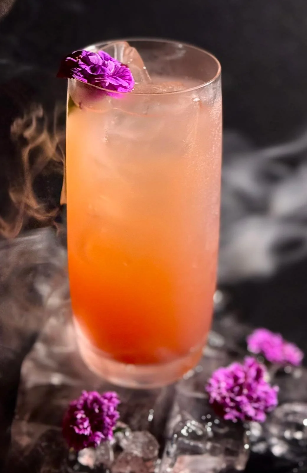 Ein bunter Cocktail in einem hohen Glas mit Eiswürfeln, verziert mit einer lila Blume. Im Hintergrund dunkler Stoff, mit einigen lila Blumen daneben.