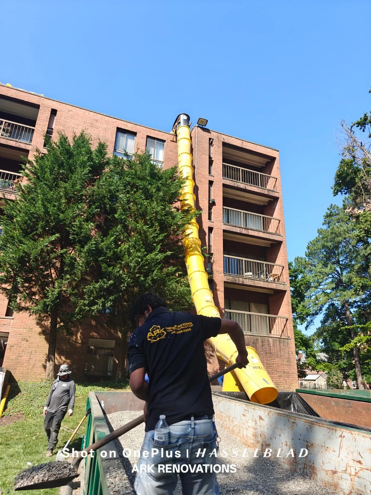 Construction workers installing a yellow vent pipe on the exterior of a brick apartment building with balconies, under a clear blue sky, with trees nearby.