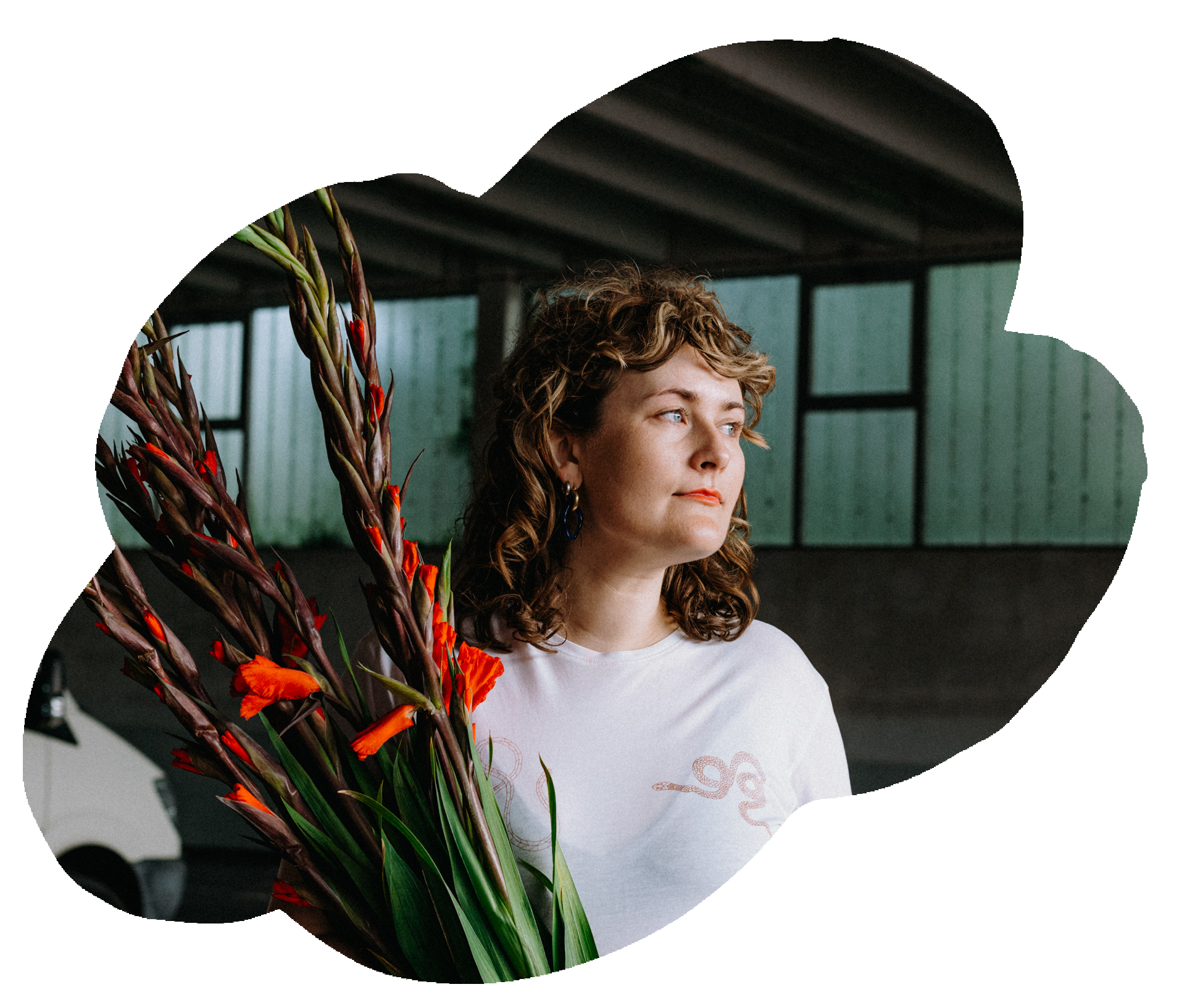 Eine Frau mit lockigem braunem Haar steht neben einem großen Blumenstrauß aus Gladiolen, in einem industrialen Raum mit grünen Wänden und großen Fenstern.