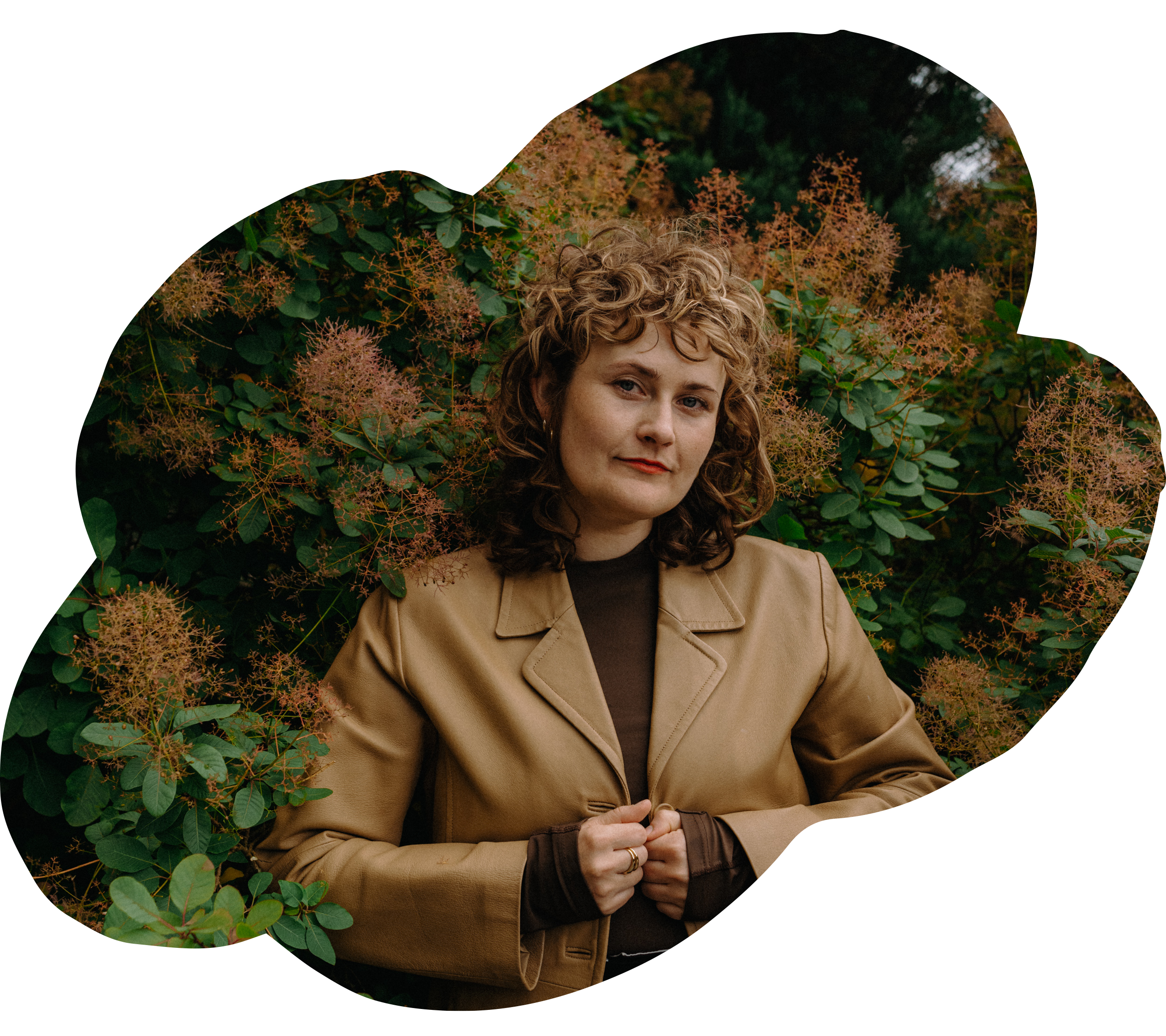 Eine Frau mit lockigem Haar trägt eine braune Jacke und steht vor einer grünen Blätterbush, umgeben von blühenden Pflanzen.