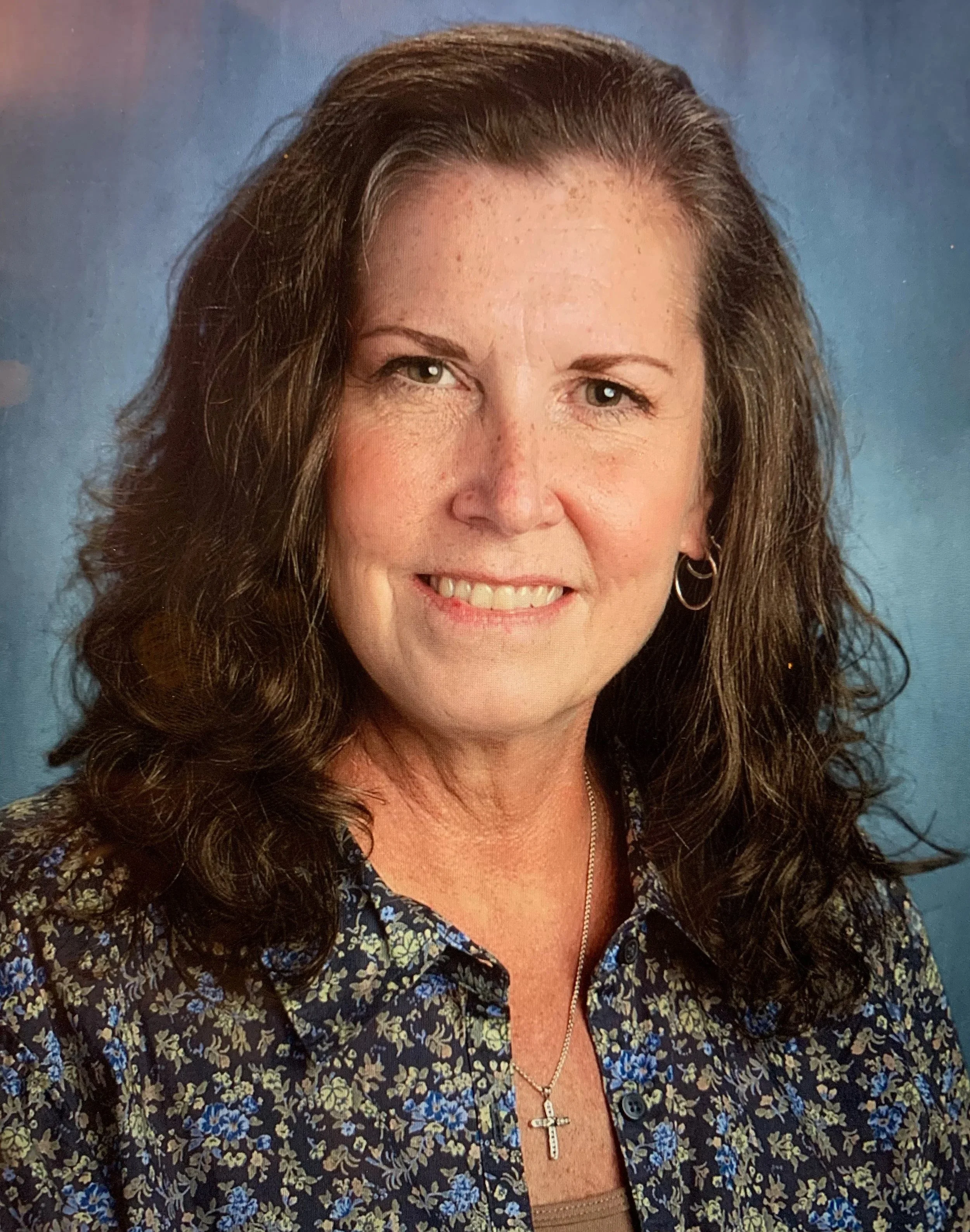 A woman with wavy brown hair, wearing a floral patterned blouse, hoop earrings, and a cross necklace, smiling in front of a blue background. Author and illustrator, Diane Fanucci.  Writer of this blog.