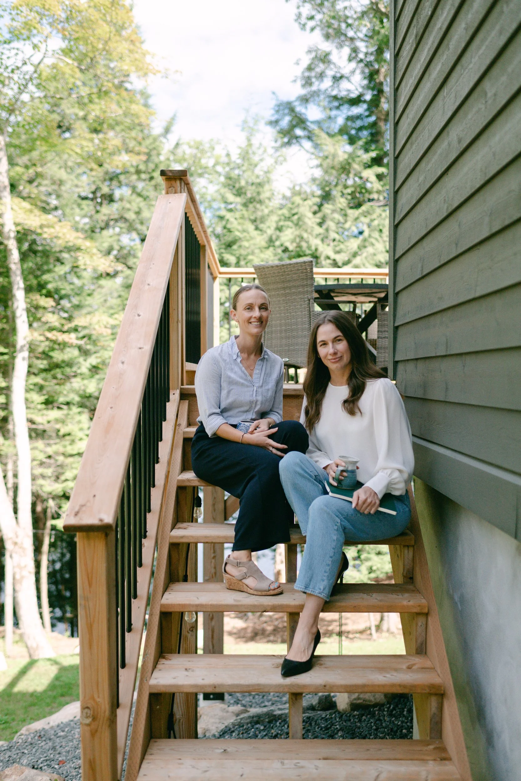 Two women sitting on wooden stairs outside, smiling, surrounded by green trees, holding mugs and a notebook.