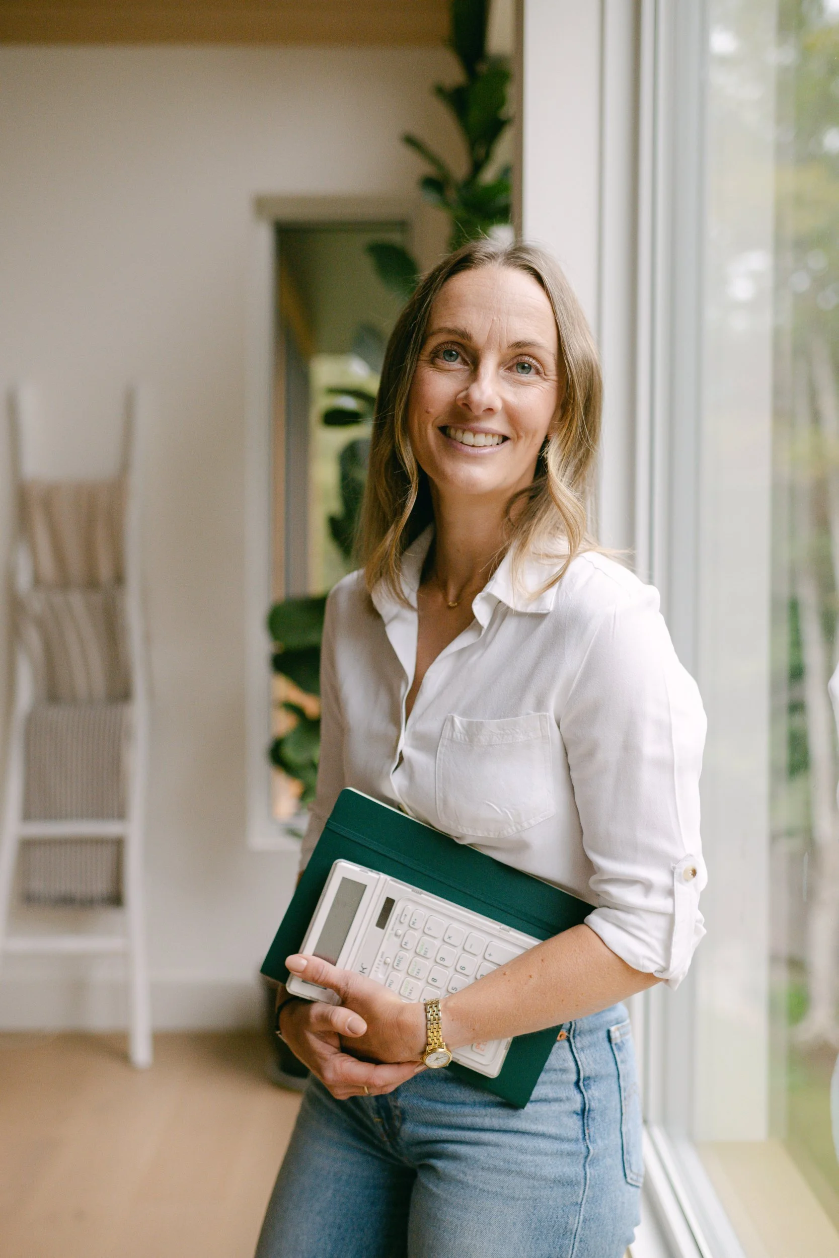 A woman standing indoors near a window, holding papers and a calculator, smiling at the camera, wearing a white blouse, blue jeans, and a gold watch.