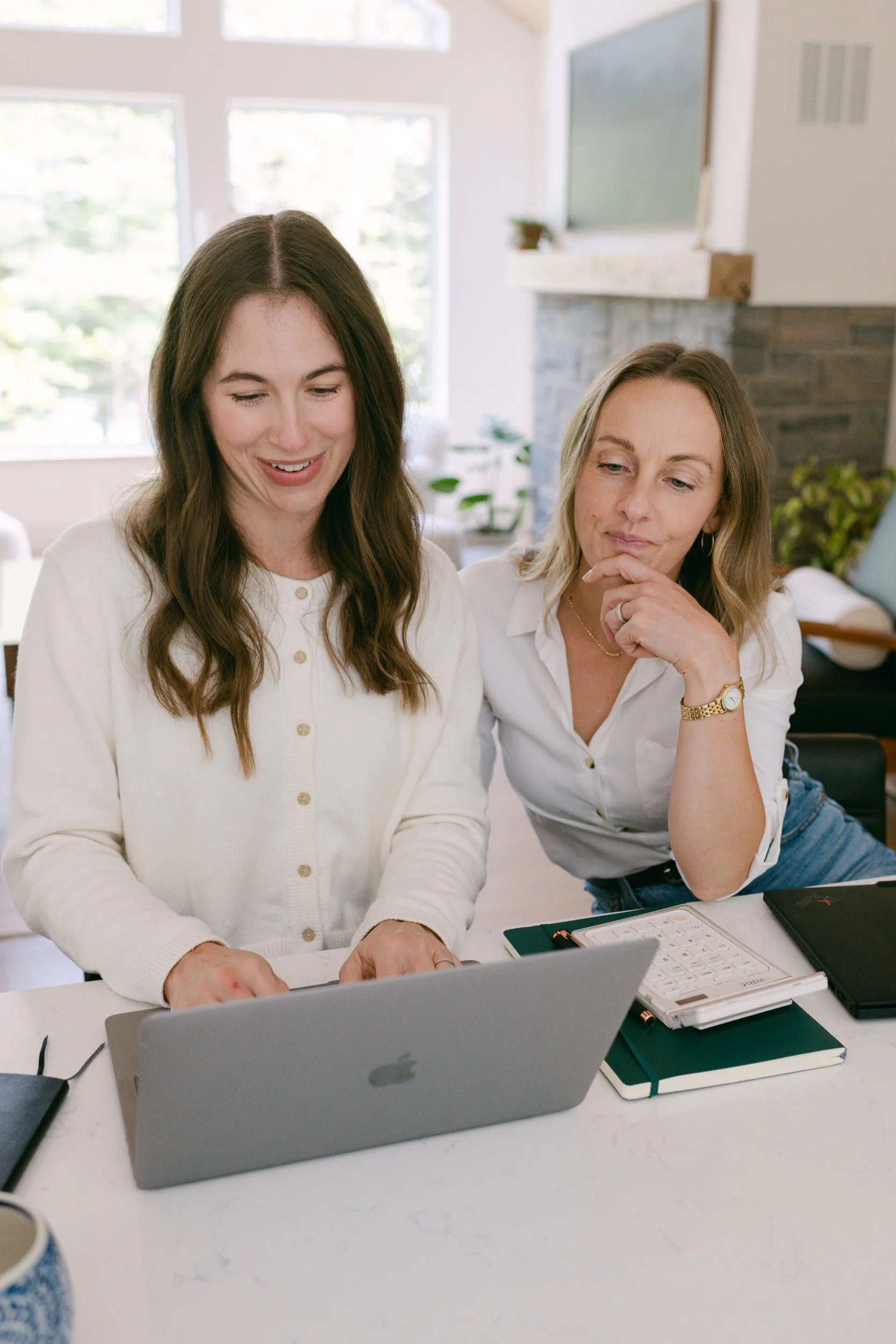 Two women sitting at a desk working on a laptop, with notebooks, a calculator, and a pen in front of them. One woman is showing something on her laptop to the other, both appearing focused.