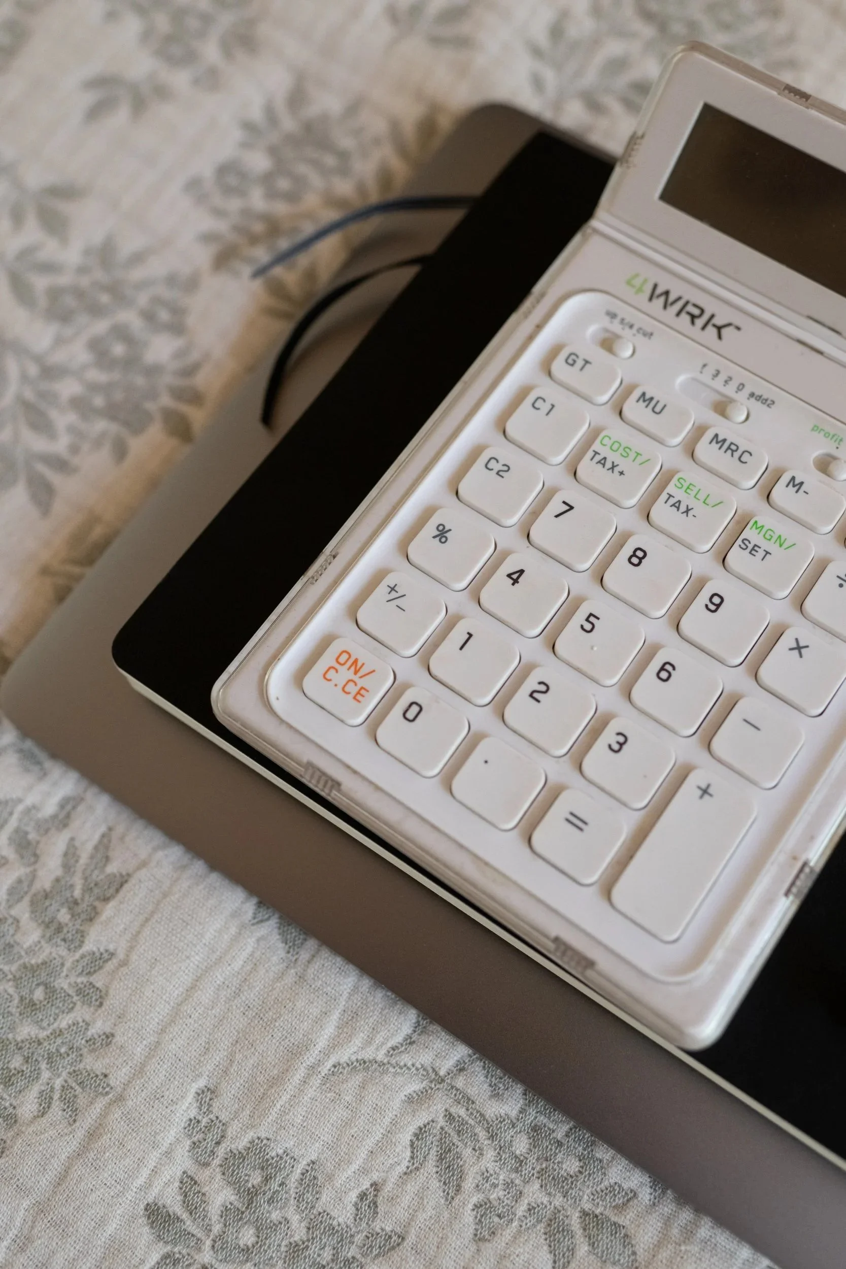 A white calculator with green and black buttons on a beige lace tablecloth.