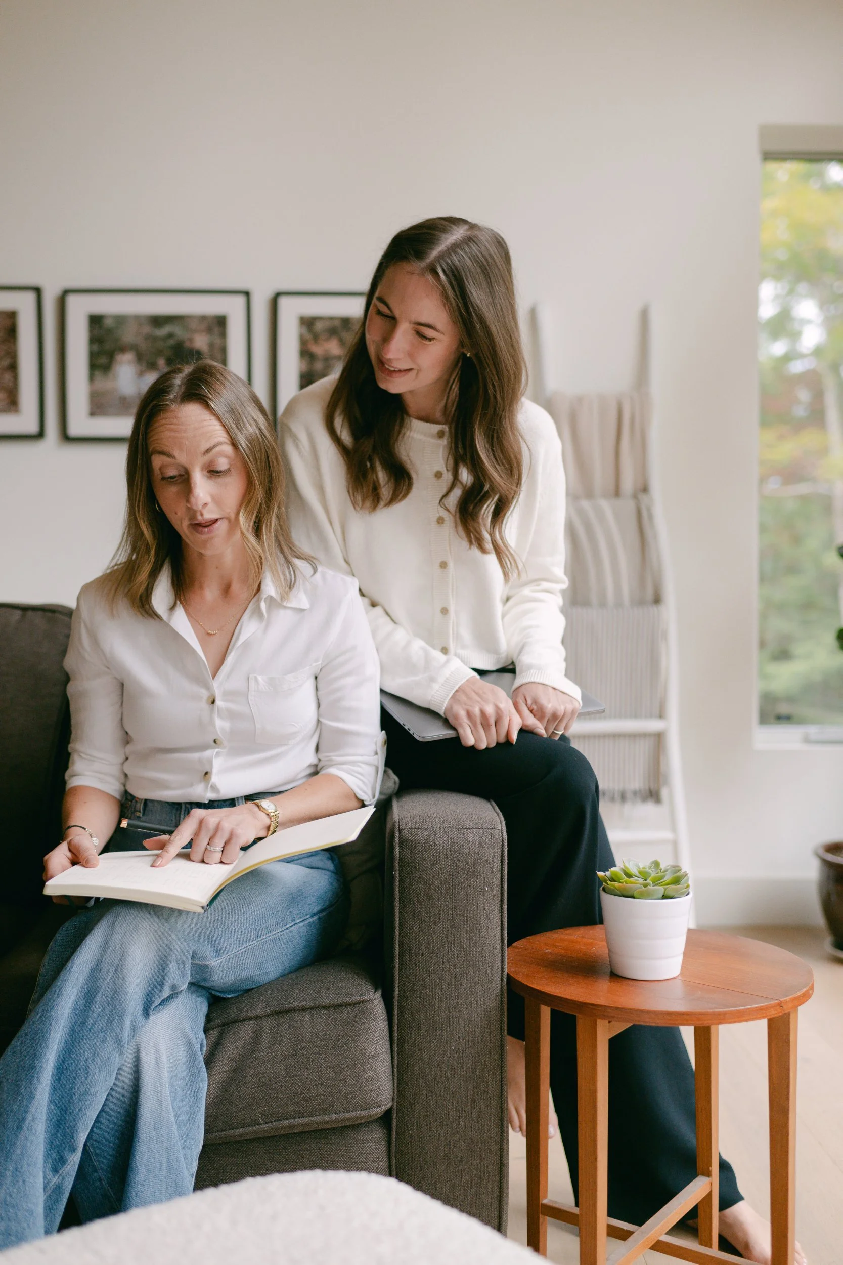 Two women sitting on a gray couch in a bright living room, one is holding a book and a tablet while the other stands nearby smiling. A small wooden table with a potted succulent is next to them.