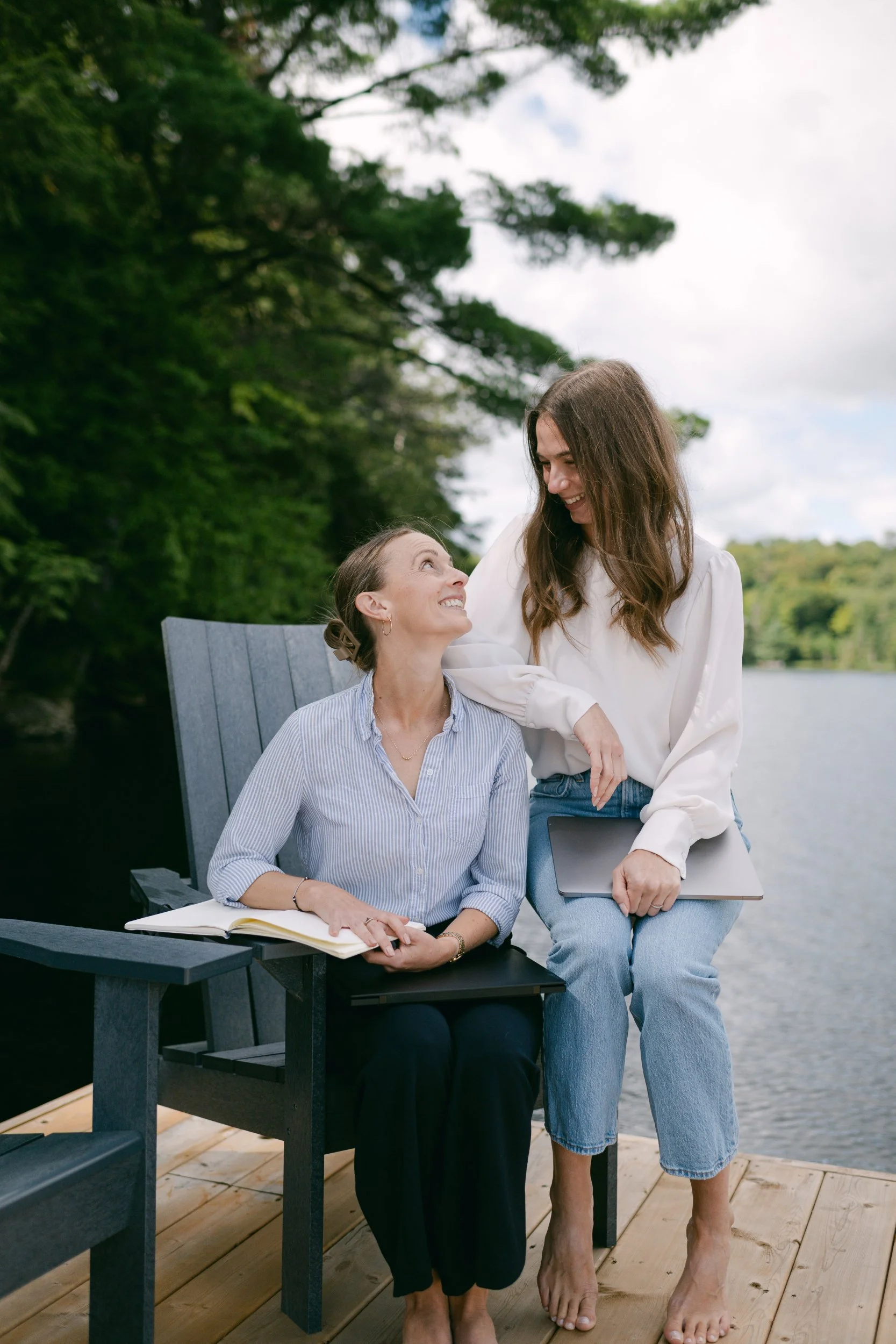 Two women, one sitting on a bench with an open notebook, and the other standing with a closed laptop, smiling at each other outdoors near a lake with trees in the background.