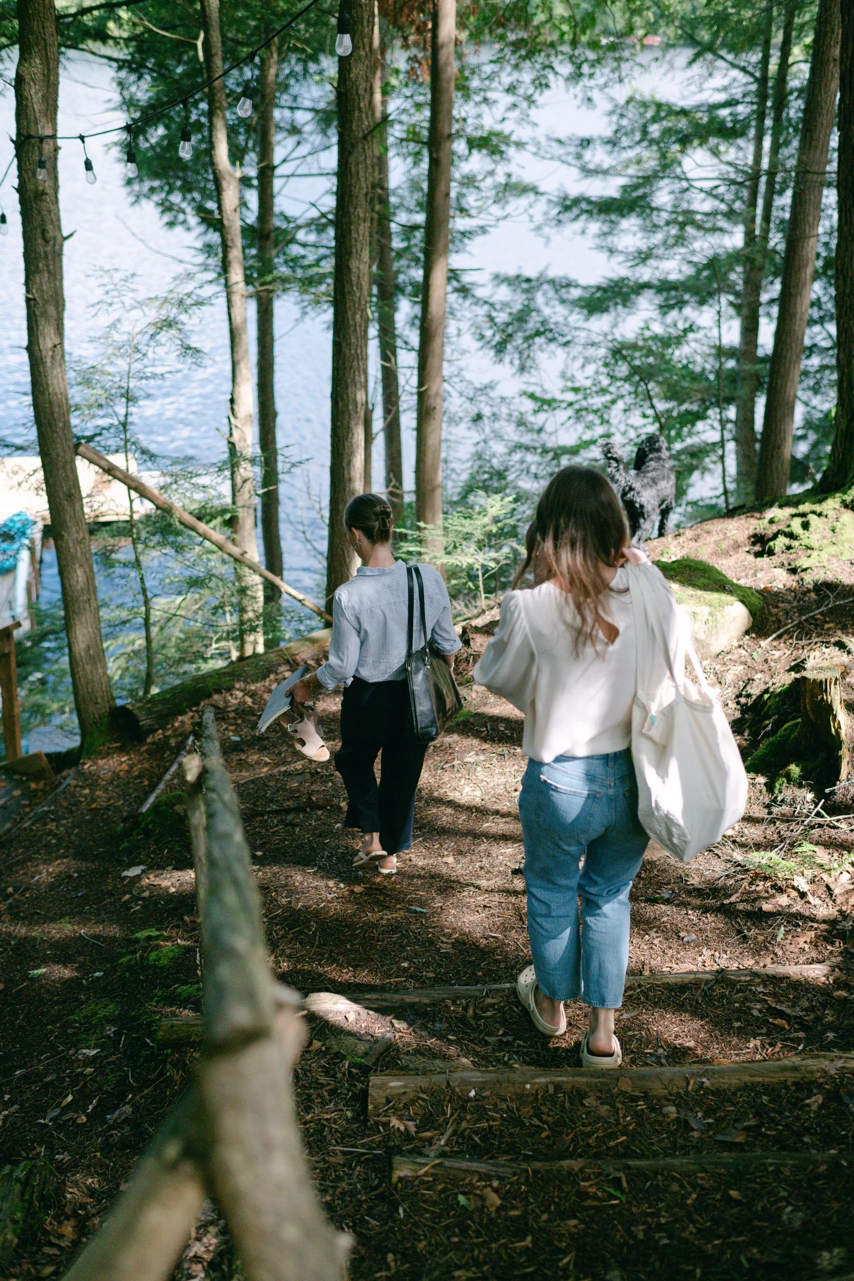 Two women walking on a dirt trail in a forested area near a lake, with tall trees and string lights hanging overhead.