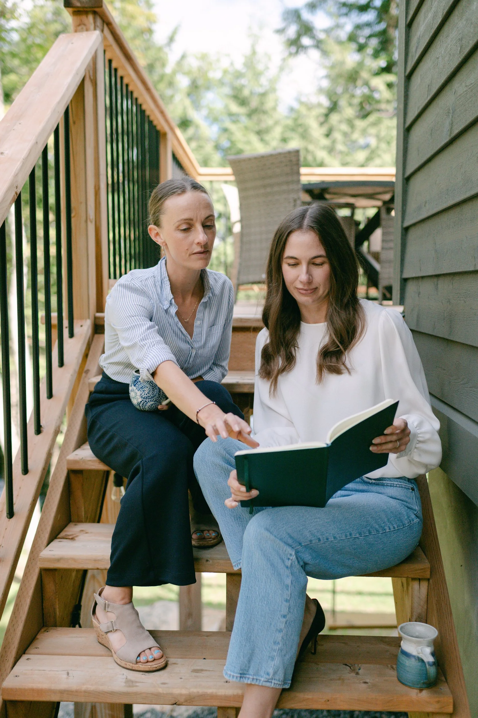 Two women are sitting on wooden outdoor stairs, looking at a book together, with trees in the background.