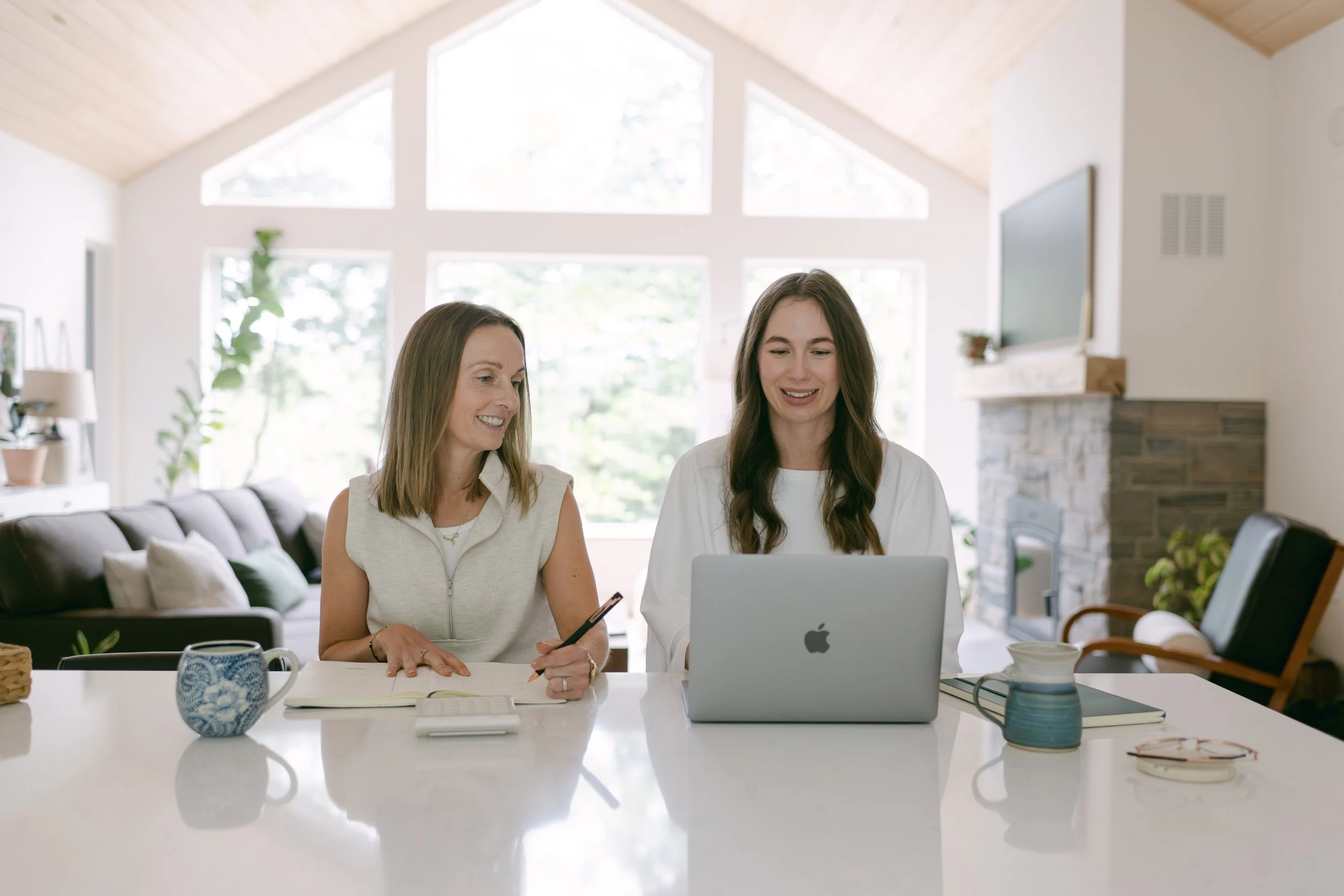 Two women sitting at a kitchen table, looking at a laptop, with notebooks, pens, and cups on the table in a bright, spacious room with large windows and a vaulted ceiling.