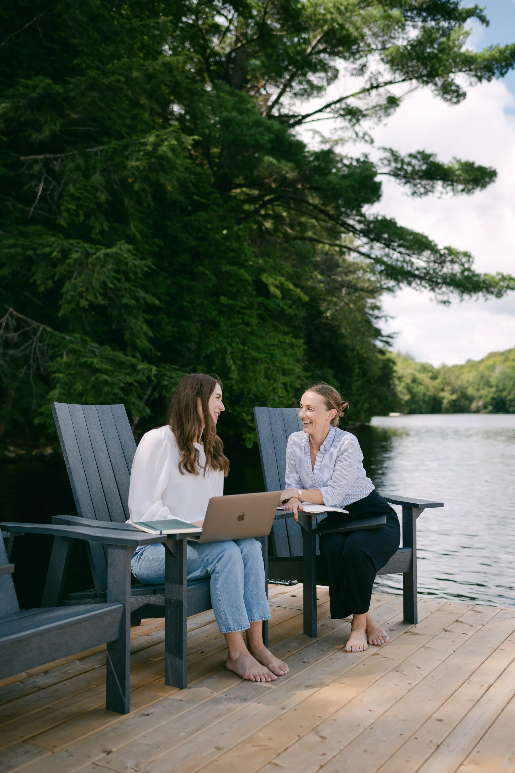 Two women sitting and talking on a wooden dock by the water, surrounded by greenery, with a laptop and books on a table nearby.
