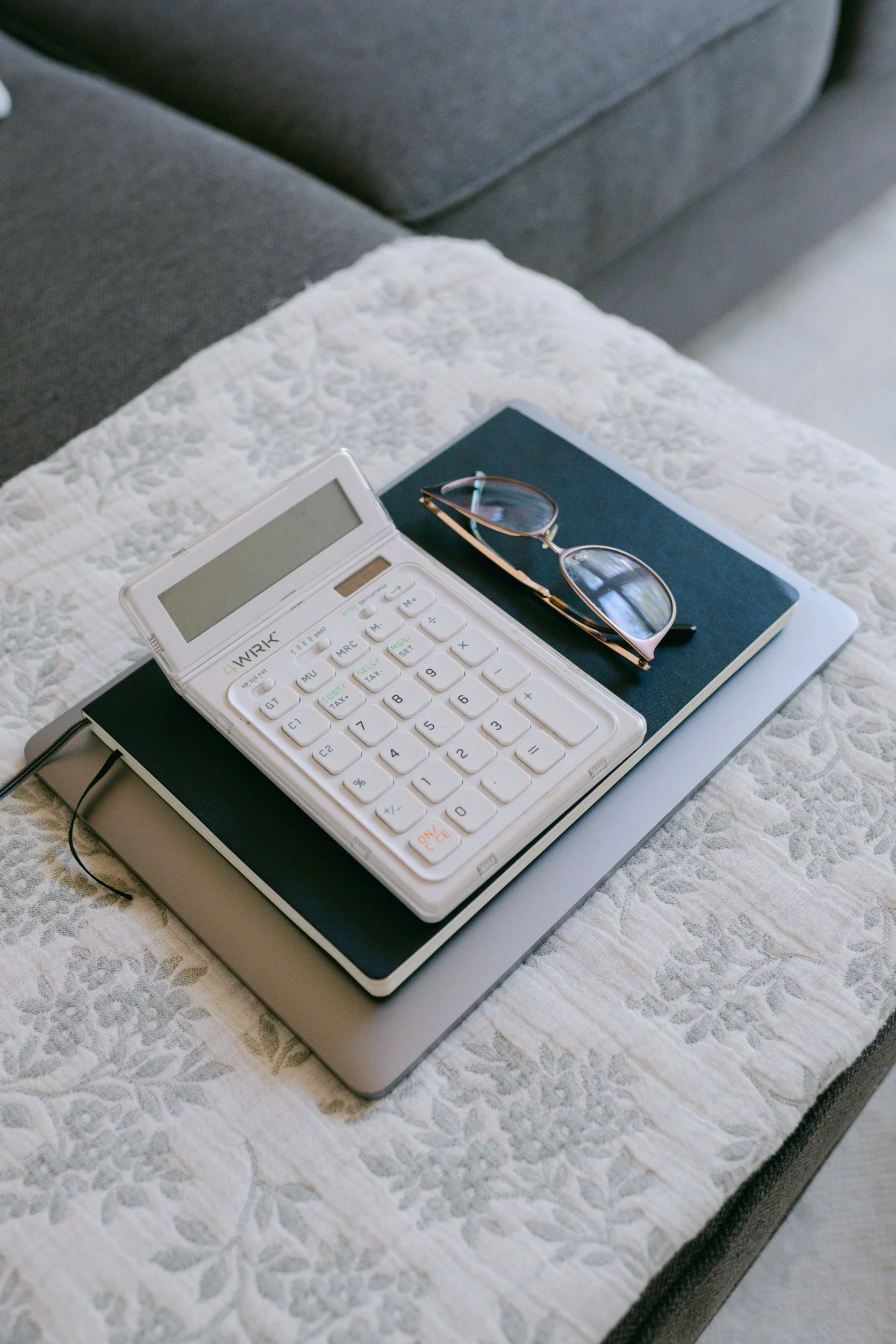 A white calculator, a pair of eyeglasses, and a closed dark blue notebook resting on a closed laptop on top of a white textured bedspread.