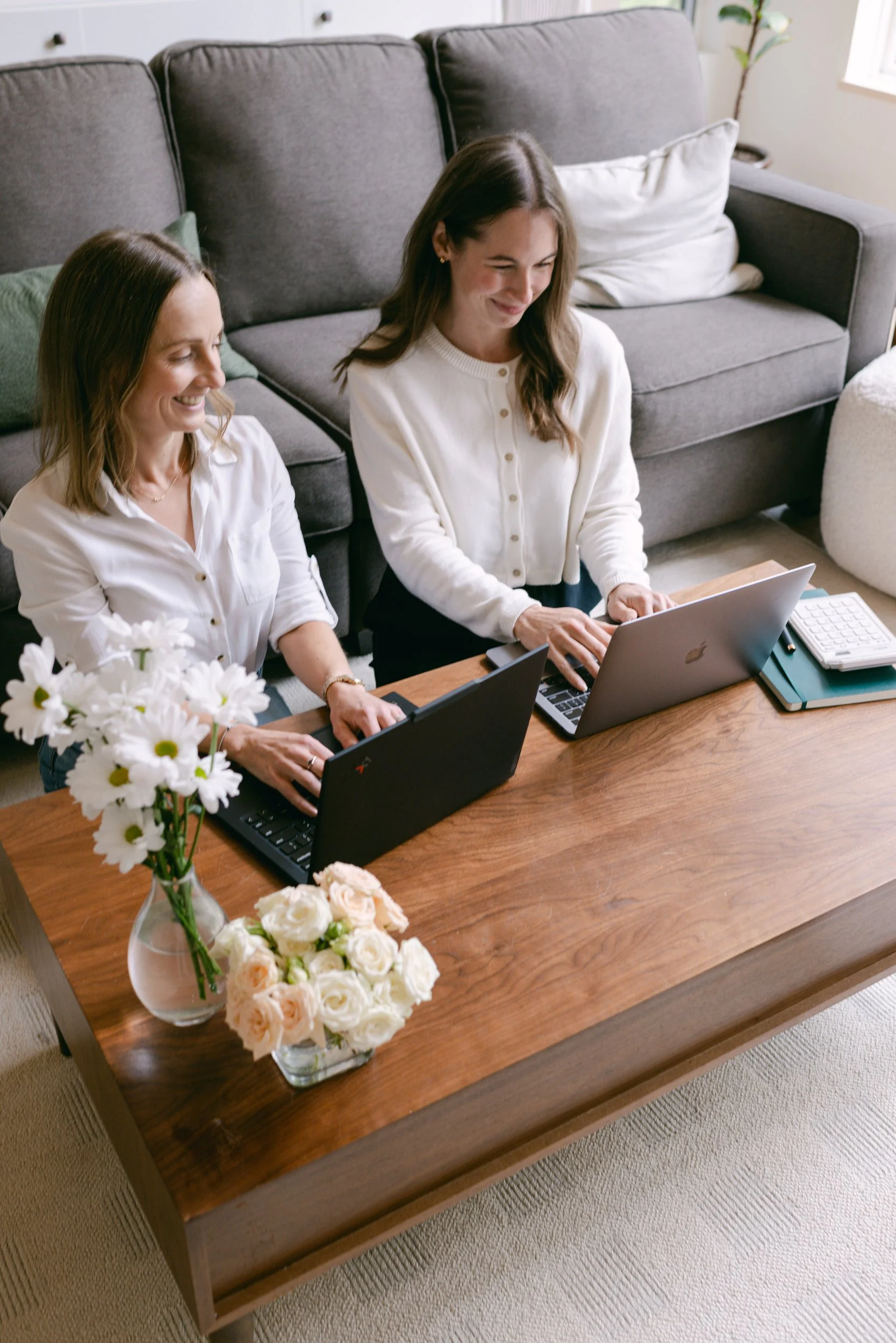 Two women sitting at a wooden table working on laptops. The table has a vase of white and peach flowers. They are in a bright living room with a gray sofa, white cushions, and a plant in the background.