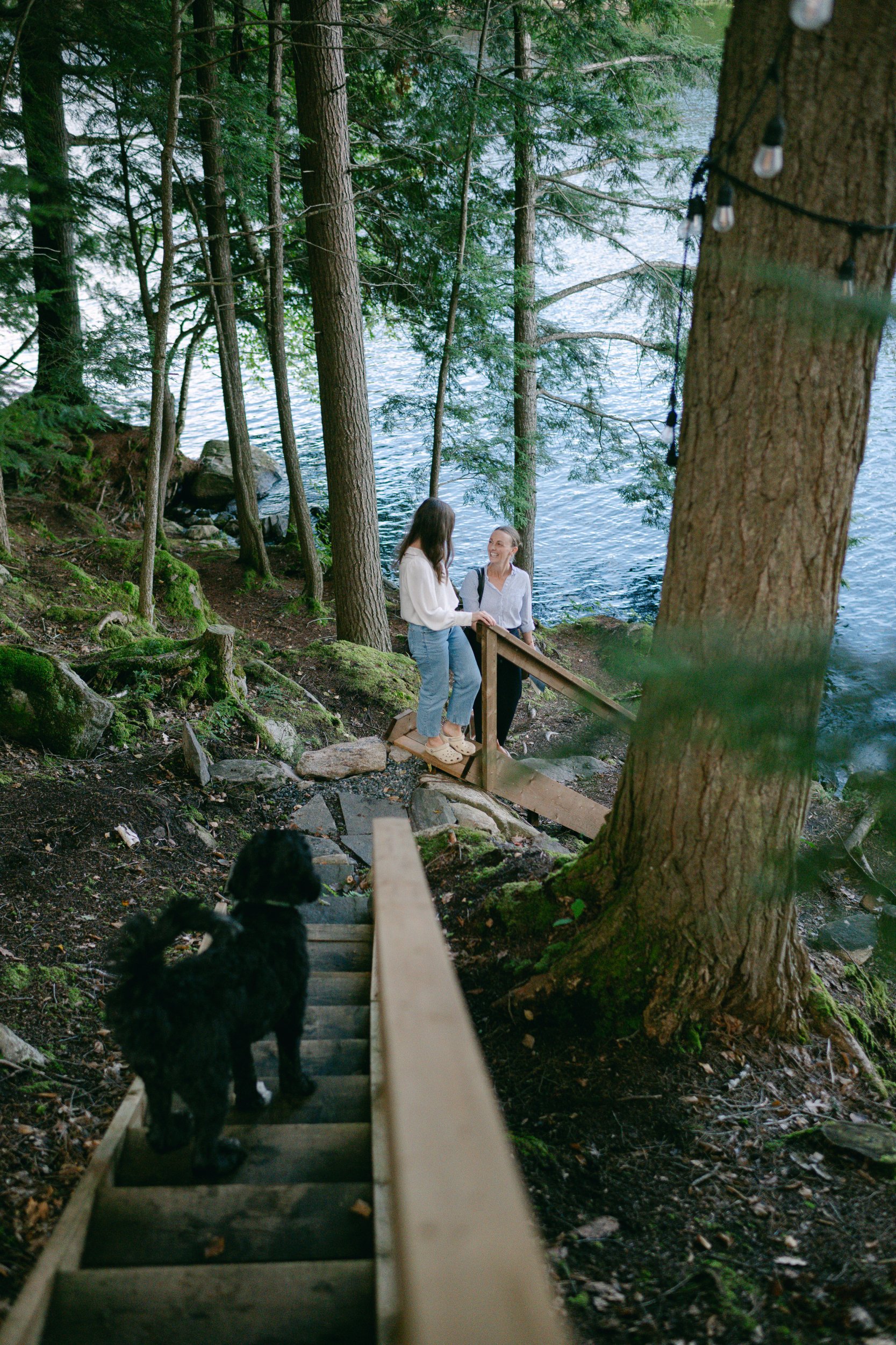 Two women standing on a wooden trail near a lake, surrounded by trees, with a black dog at the bottom of a wooden staircase.
