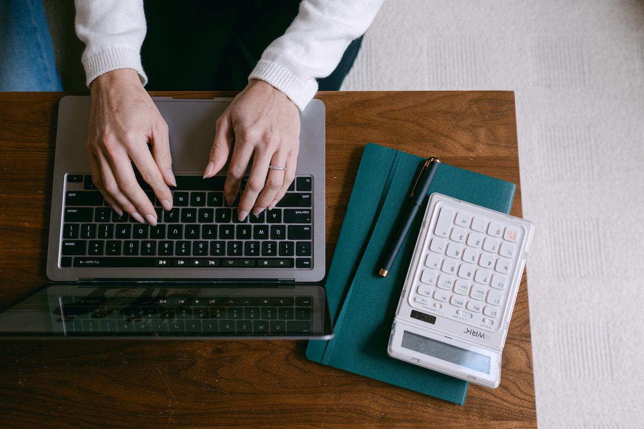 Person working on laptop at a wooden desk with notebooks, a pen, and a calculator.