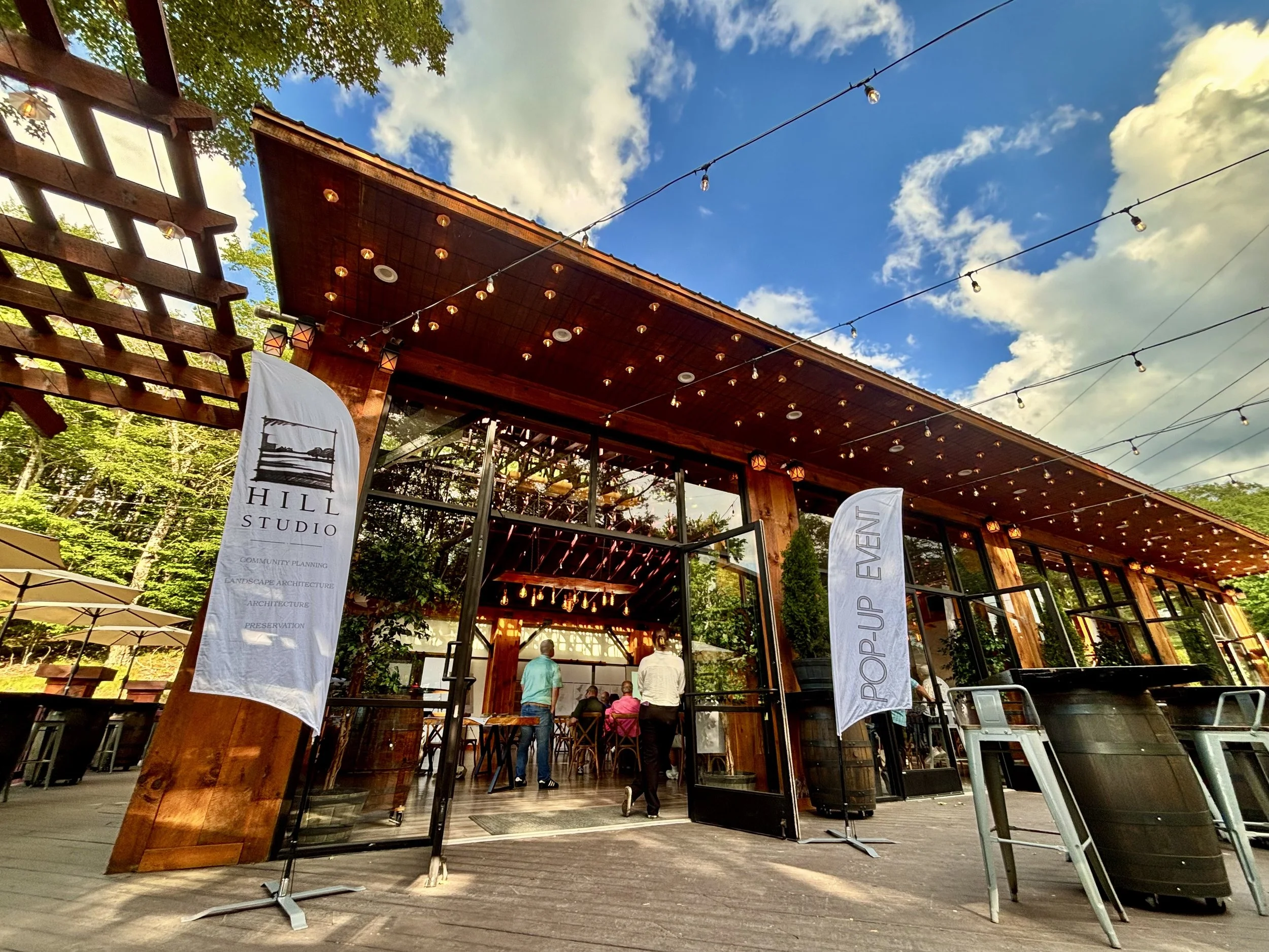 An outdoor venue with a wooden structure, string lights, and banners displaying text about community planning and architecture. People are gathered inside, with trees and a blue sky with clouds visible in the background.