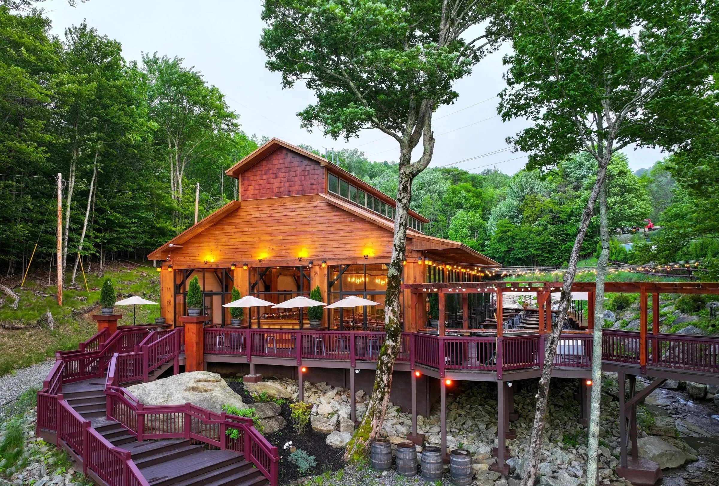 A wooden restaurant or house with a large outdoor deck, string lights, surrounded by trees and greenery, with a staircase leading down to a rocky area.