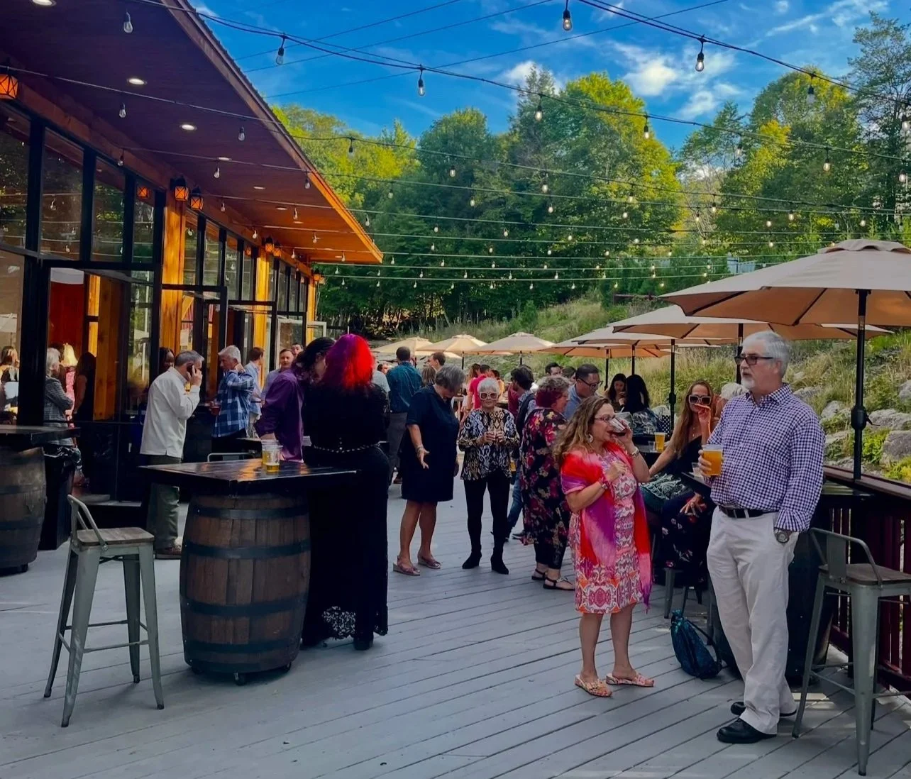 People enjoying an outdoor gathering at a patio with string lights, umbrellas, and a building with large windows, surrounded by trees.