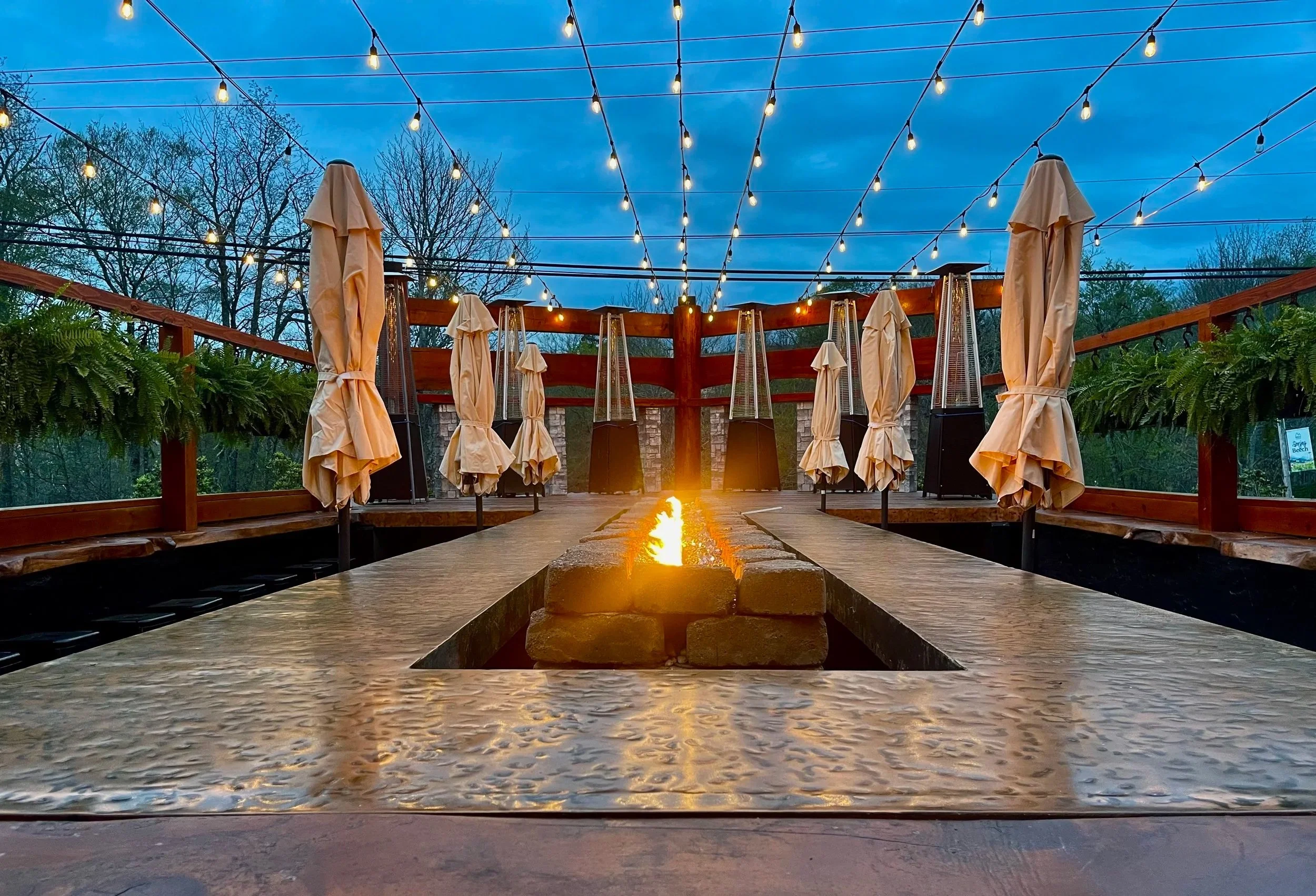 Outdoor patio with string lights overhead, beige umbrellas closed, and a central fire feature on a stone table, surrounded by greenery and a wooden railing, at dusk.
