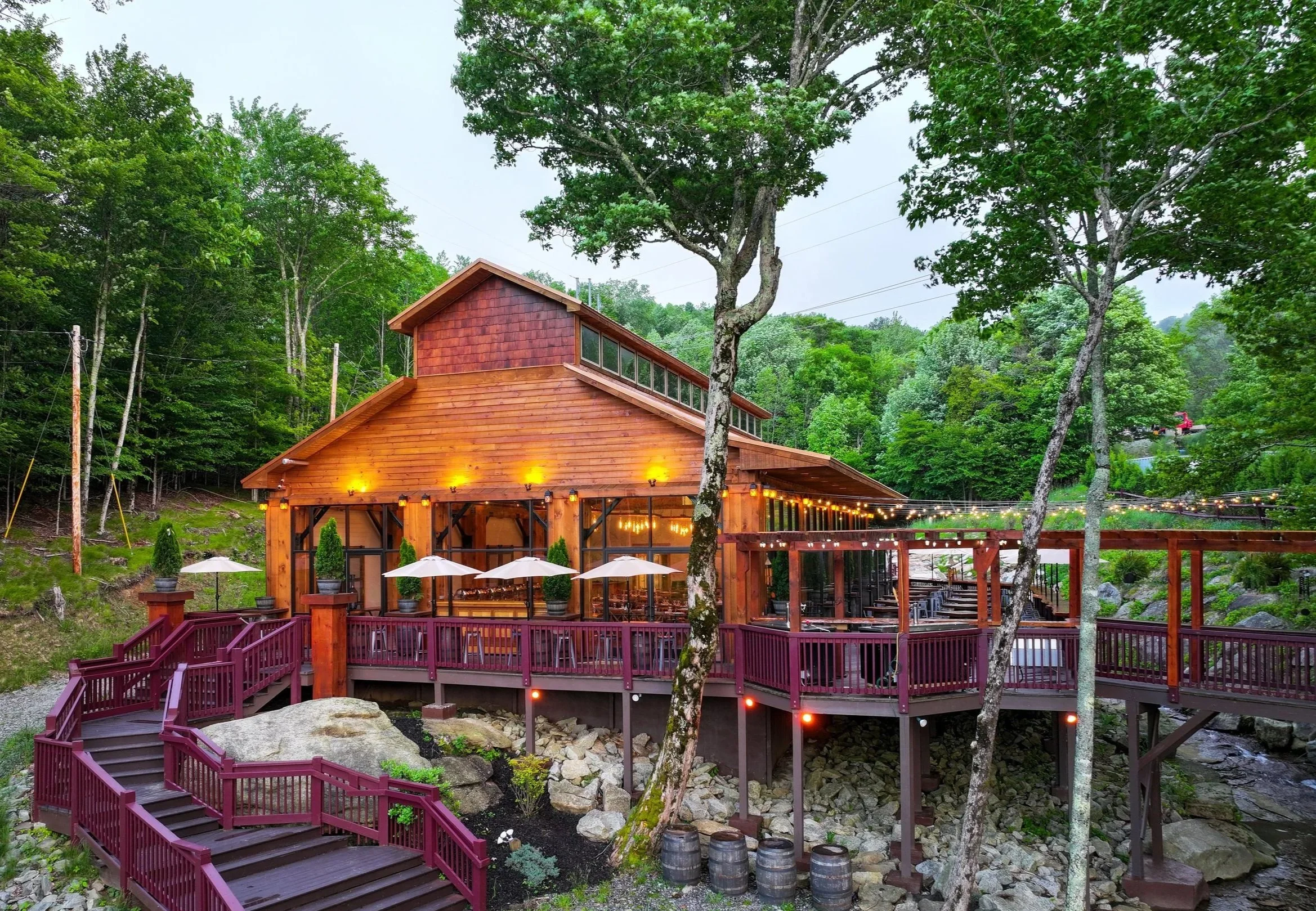 A rustic wooden building with outdoor seating, string lights, and a red deck, set among green trees.