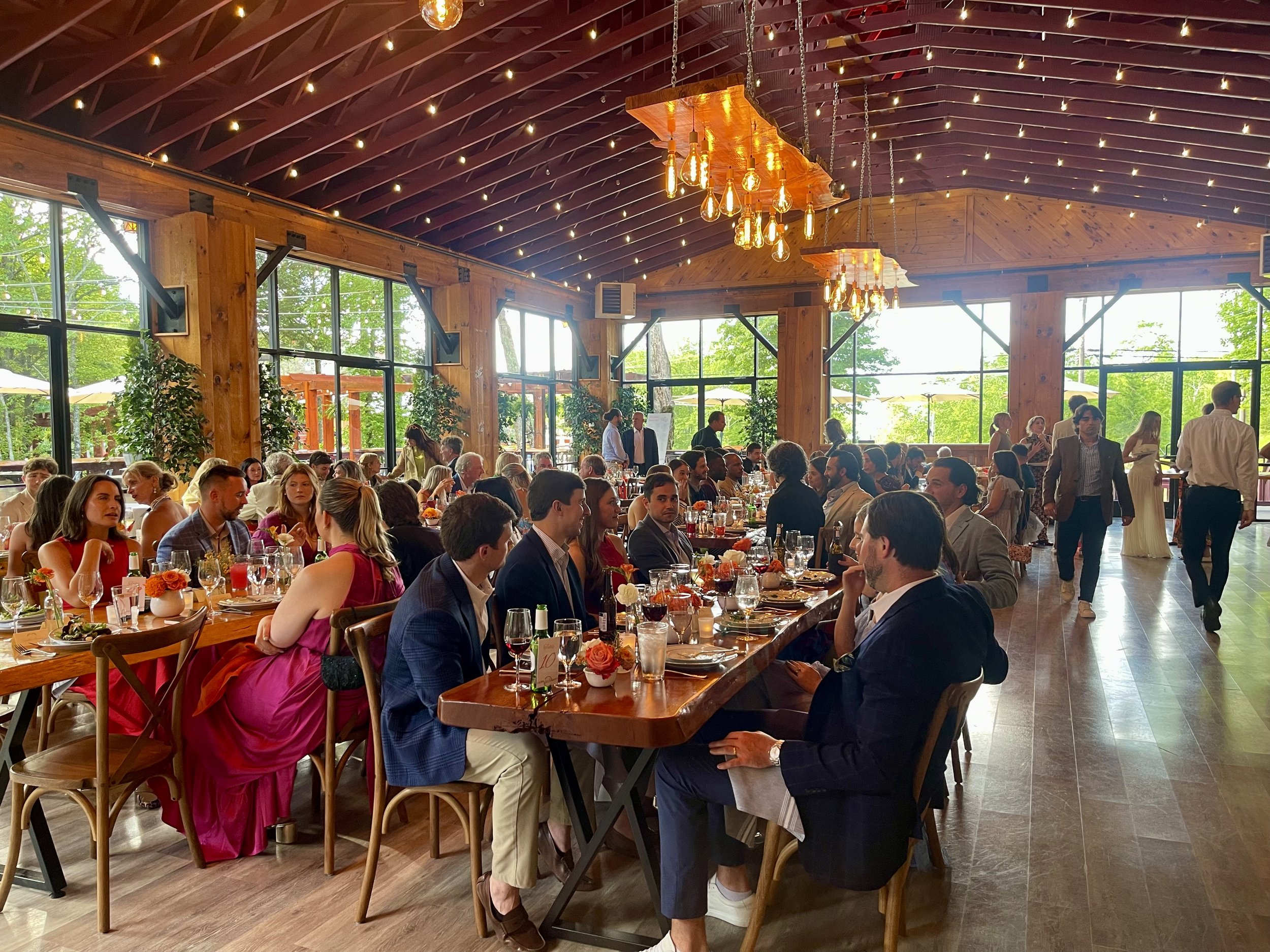 Guests seated at long tables with floral centerpieces in a spacious, rustic restaurant with wooden beams, large windows, hanging Edison bulb chandeliers, and string lights on the ceiling during daytime.
