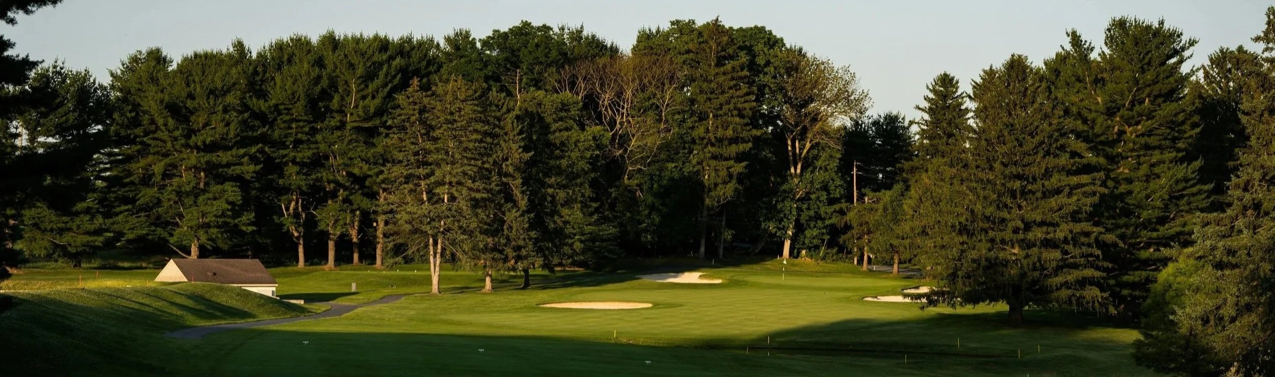 A golf course with green fairways, sand bunkers, a flagstick on the putting green, surrounded by tall trees, and a small shed or building in the background.