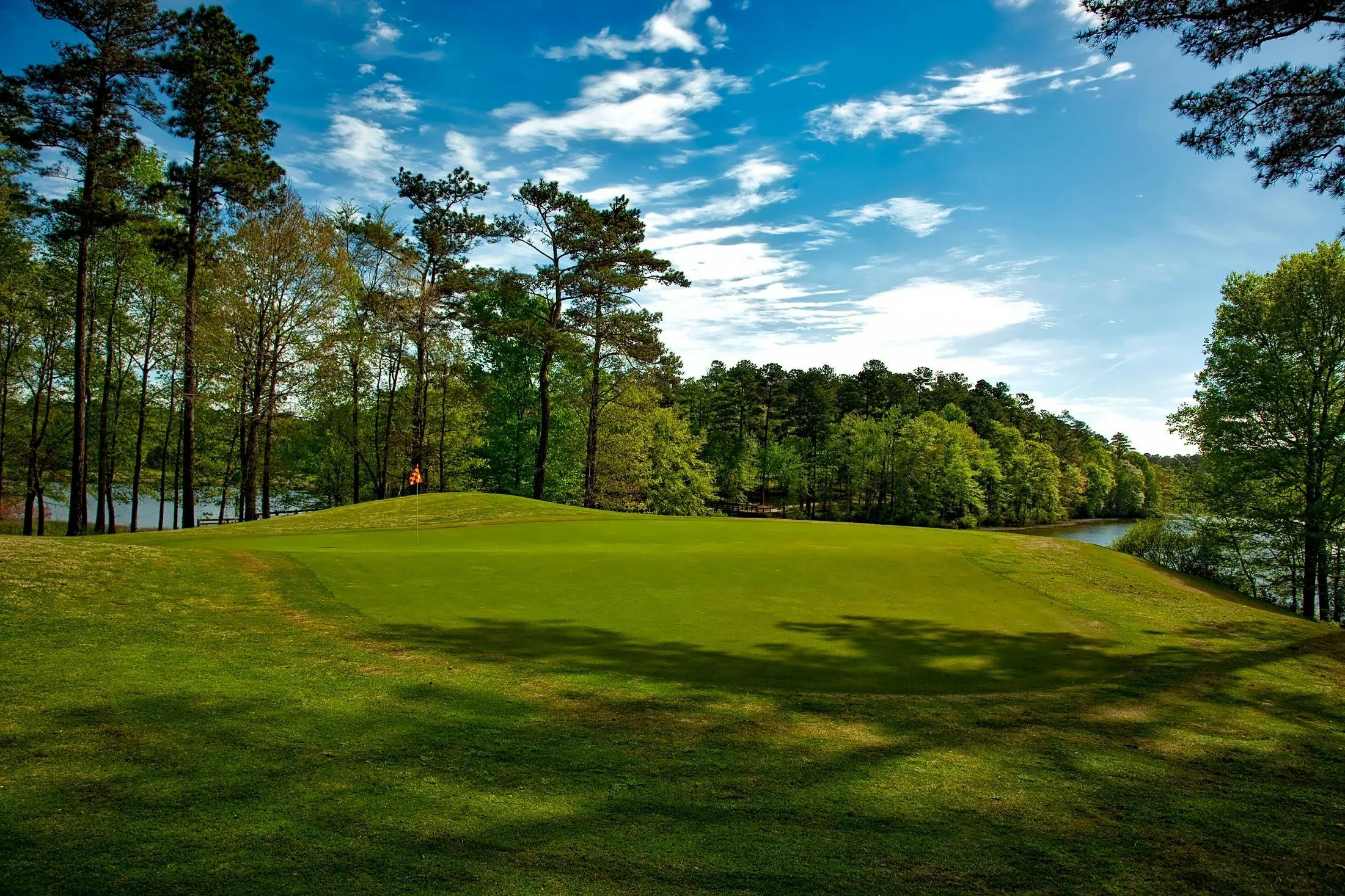 Golf course with green grass, a hole with a flag, surrounded by trees near a body of water, under a partly cloudy sky.