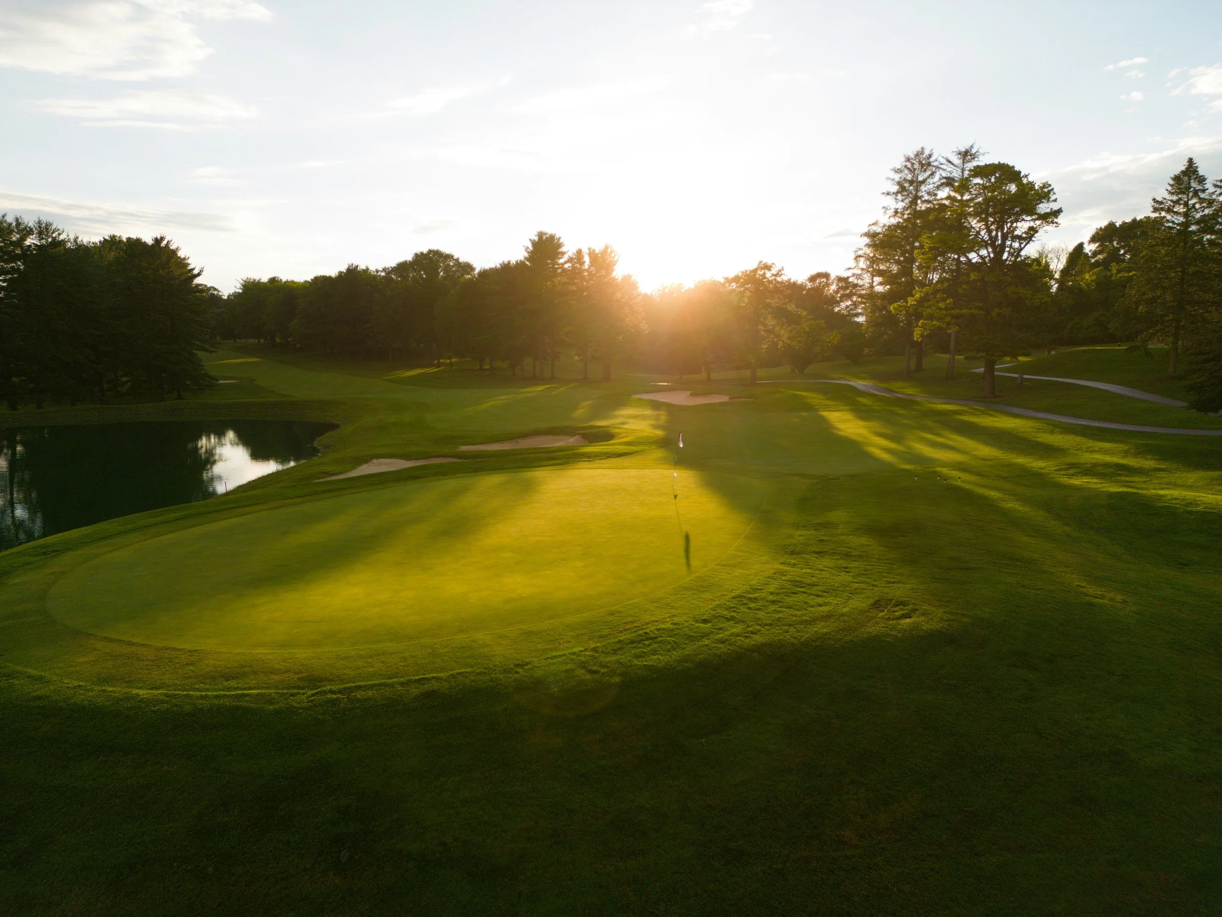 A golf course at sunset with a putting green, flag, sand traps, water hazard, and trees.