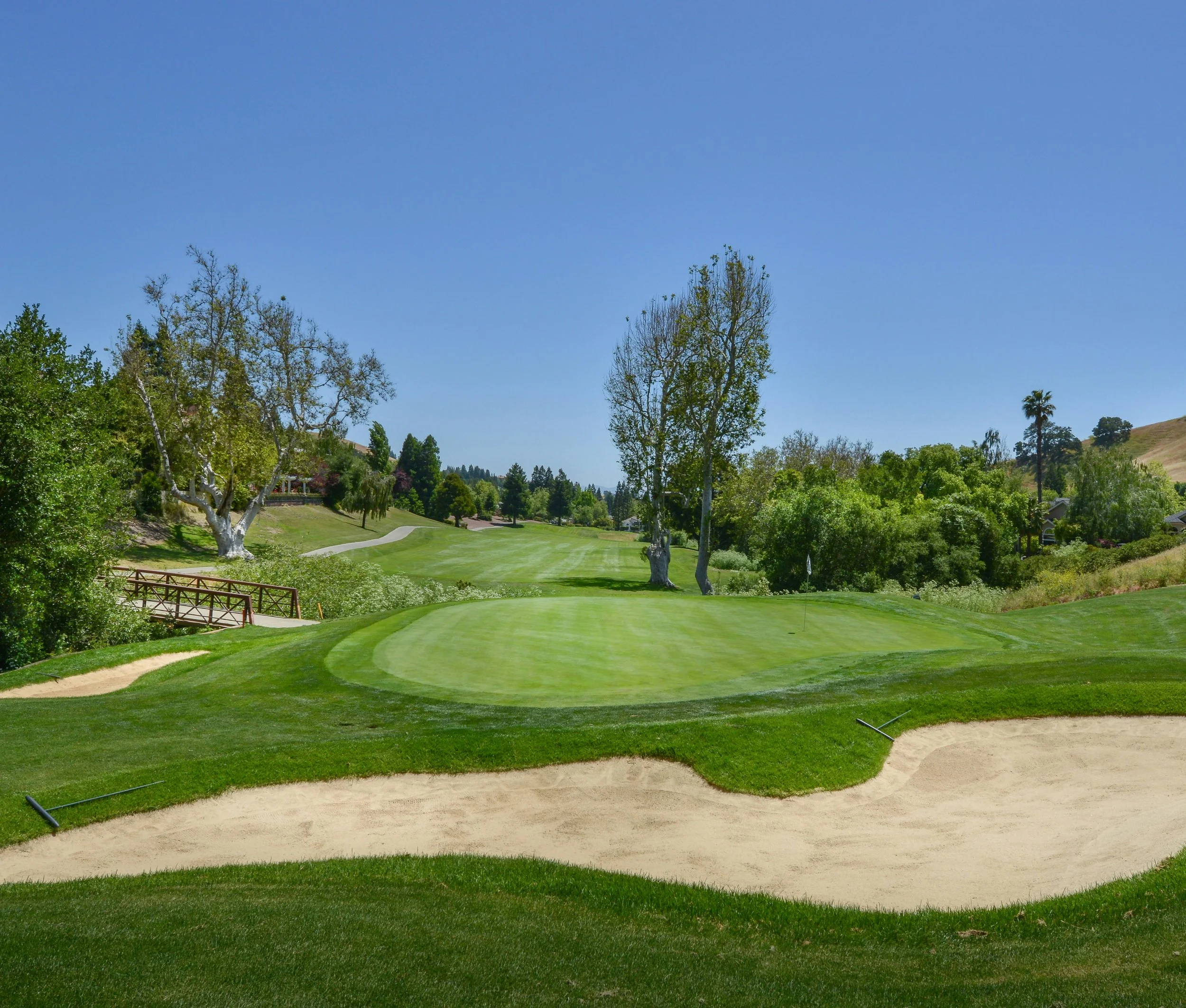 A golf course with a putting green, sand bunkers, trees, and a clear blue sky.