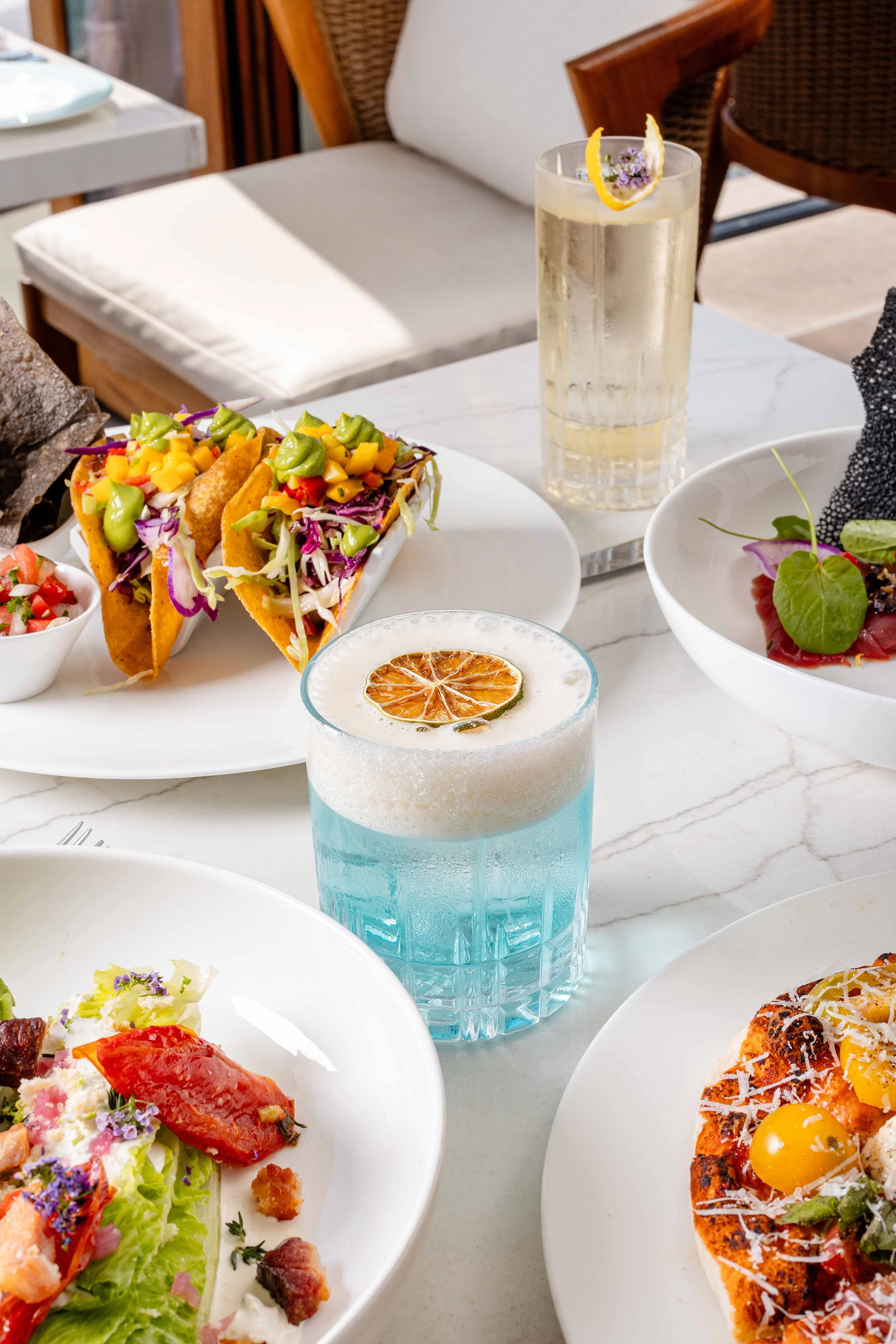 A table set with tacos, a blue cocktail with a citrus slice, a tall glass of water with lemon, a salad, and a pizza on white plates. Photo by Summerside Creative A Hotel Social Media & Creative Agency Serving Hospitality Brands