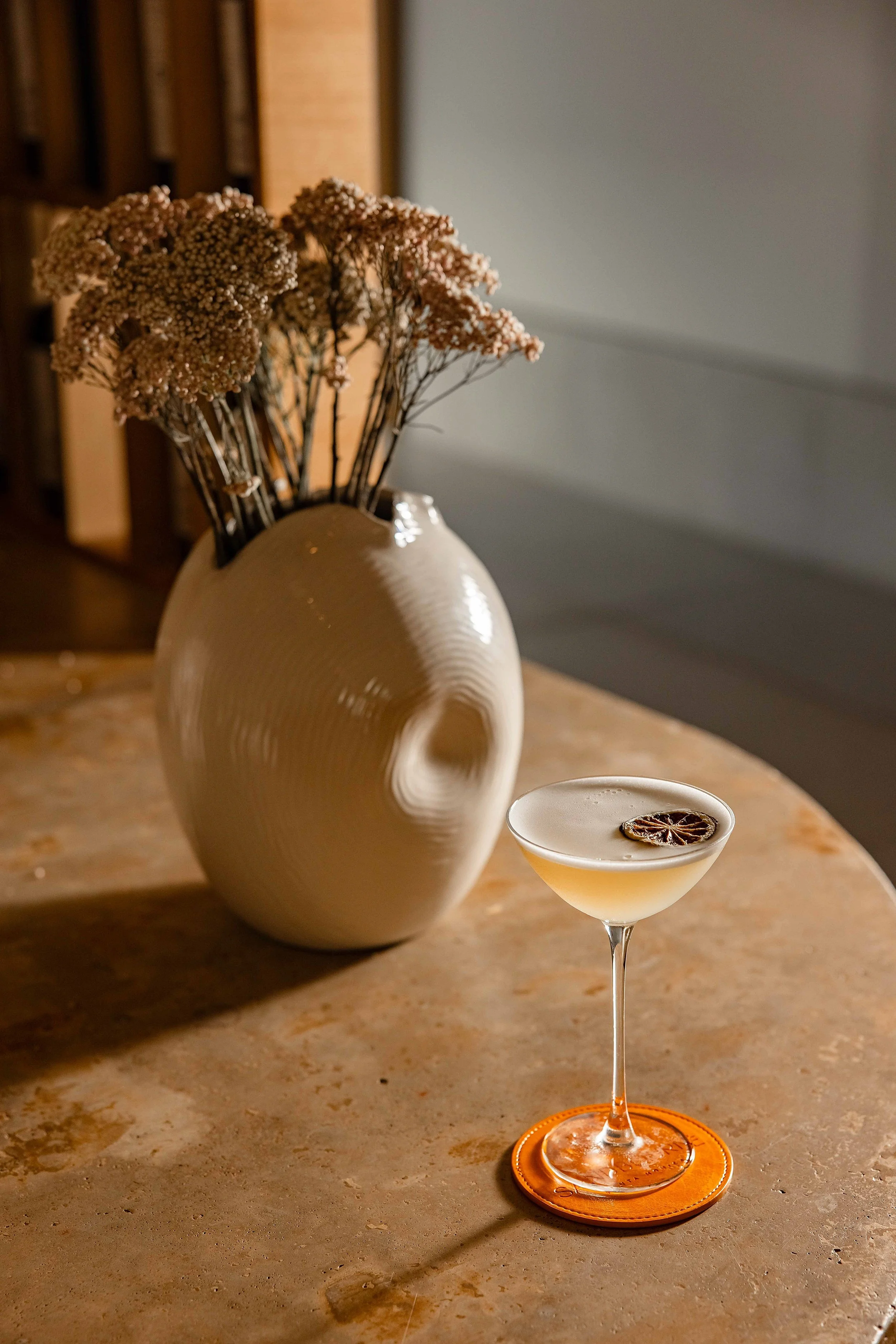 A beige ceramic vase with dried flowers and a light-colored cocktail with a dried citrus slice garnish on a marble table. Photo by Summerside Creative A Hotel Social Media & Creative Agency Serving Hospitality Brands