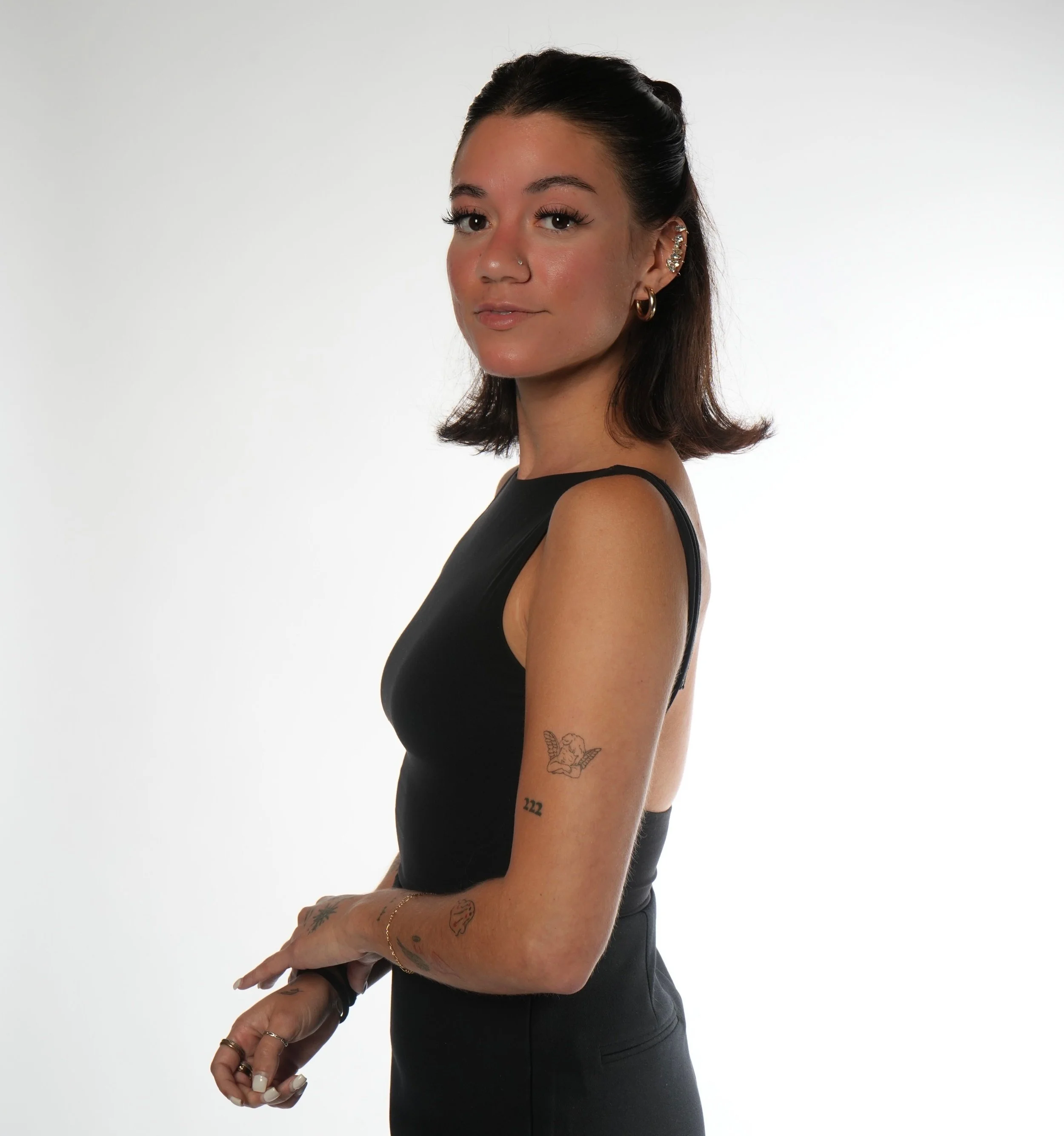A young woman with dark brown hair styled in a bob, wearing a black sleeveless top, jewelry including earrings and a nose ring, and Tattoos on her arm, posing against a plain white background.