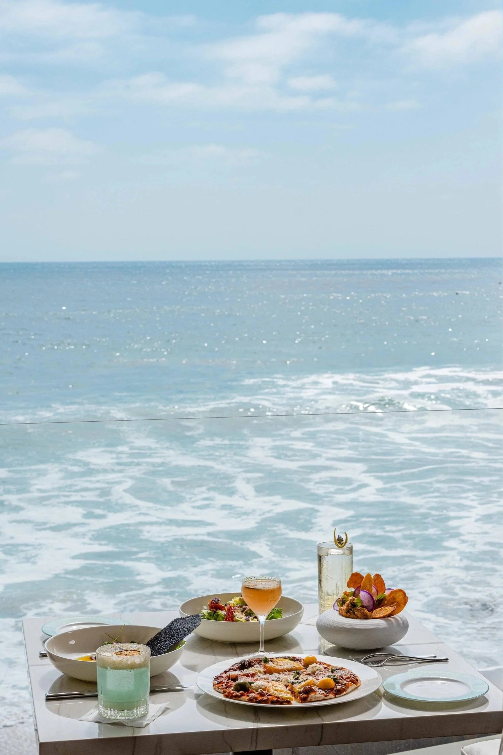 Table set with pizza, salad, drinks, and appetizers by the ocean. Photo by Summerside Creative A Hotel Social Media & Creative Agency Serving Hospitality Brands